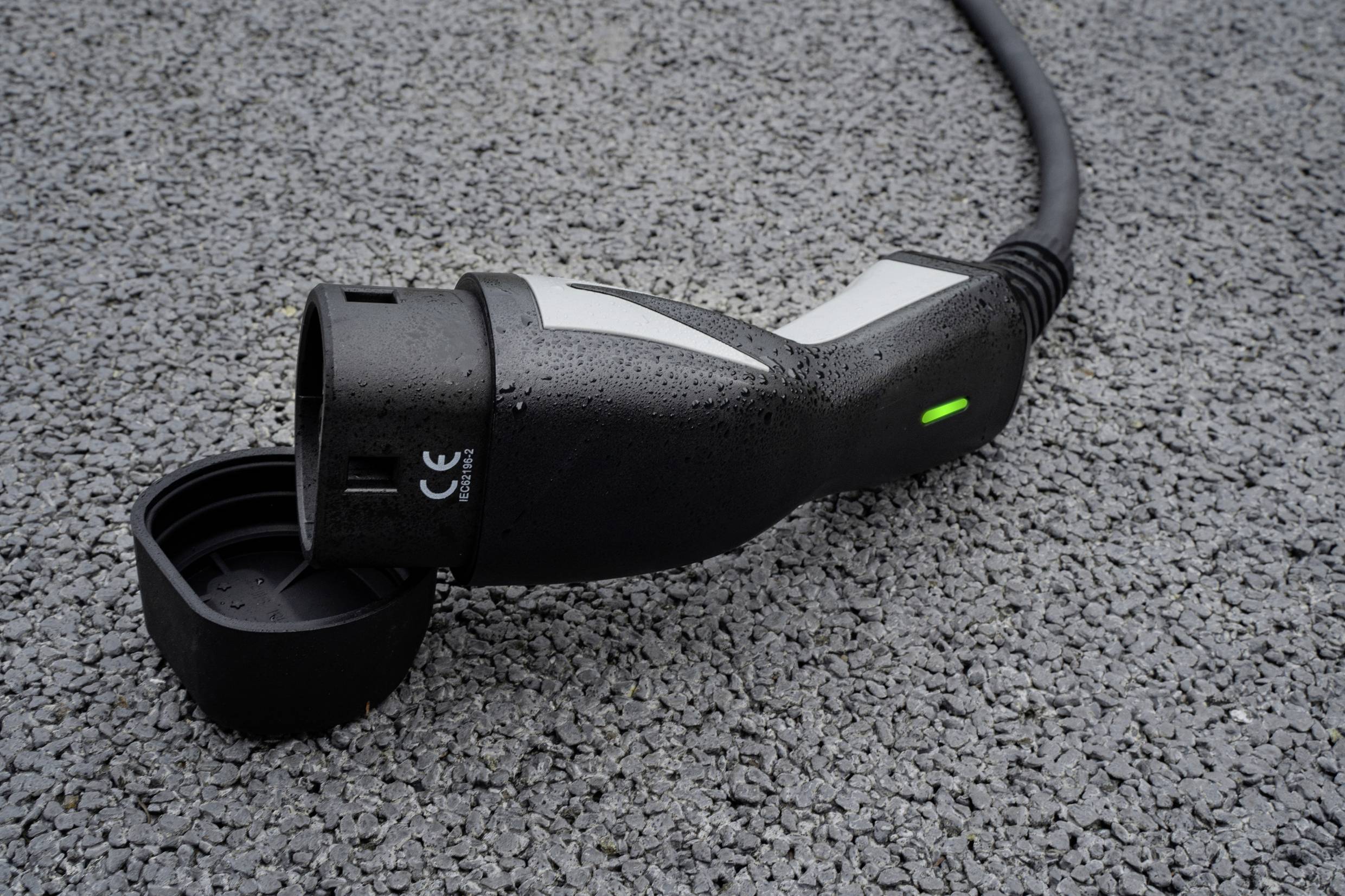Nahaufnahme eines Elektrofahrzeug-Ladesteckers auf nassem Asphalt. Ein grünes Licht leuchtet am Stecker.