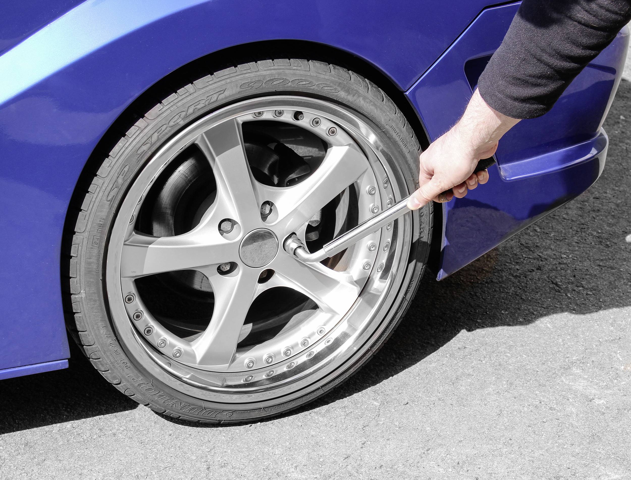 Eine Person zieht mit einem Schraubenschlüssel eine Radmutter an einem blauen Auto fest. Das Auto steht auf einer Asphaltfläche.