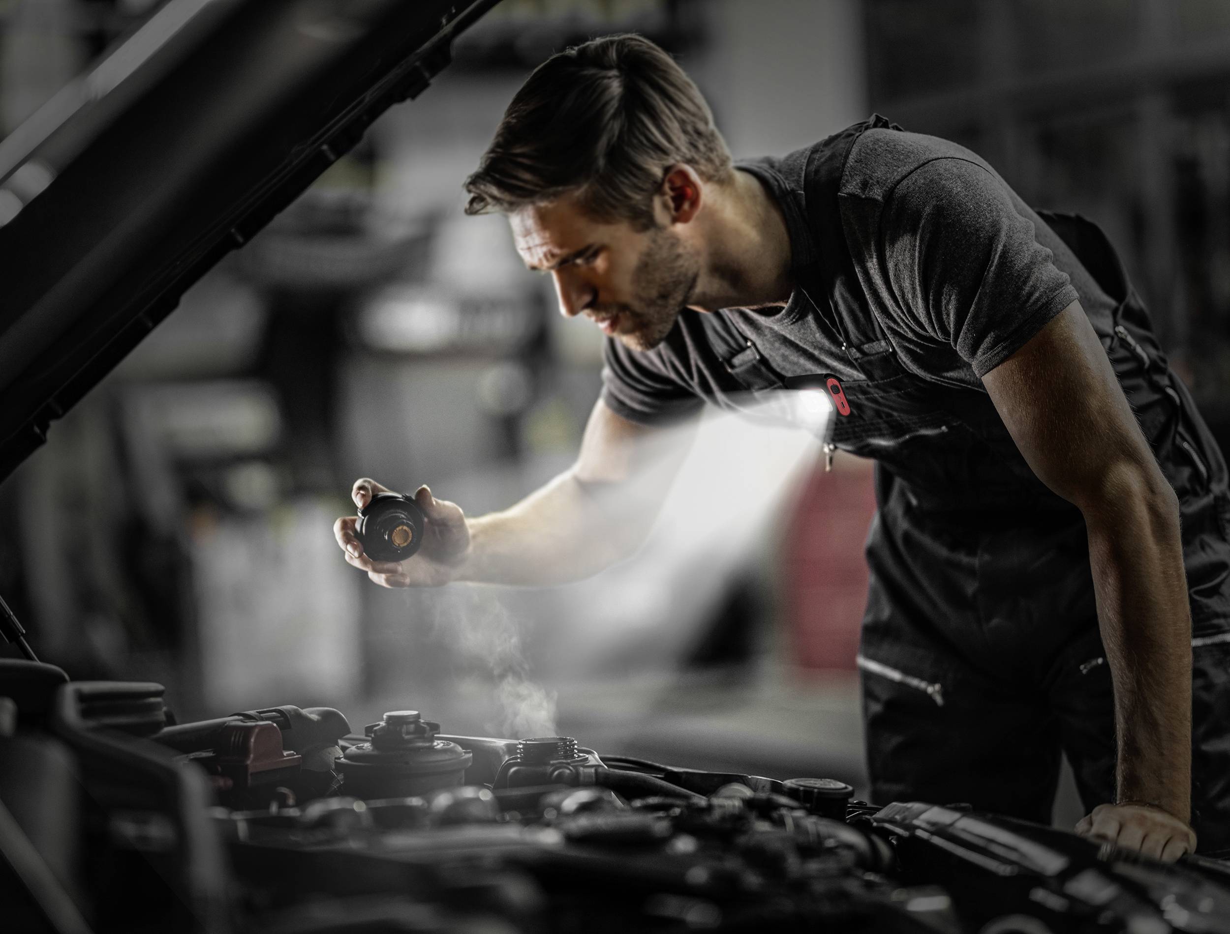 Ein Mechaniker inspiziert mit einer Taschenlampe den Motor eines Autos in einer Werkstatt. Der Fokus liegt auf der konzentrierten Arbeit.