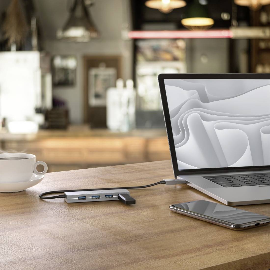Laptop auf Tisch in Café, verbunden mit USB-Hub, daneben Smartphone und Kaffeetasse. Hintergrund unscharf mit Café-Atmosphäre.