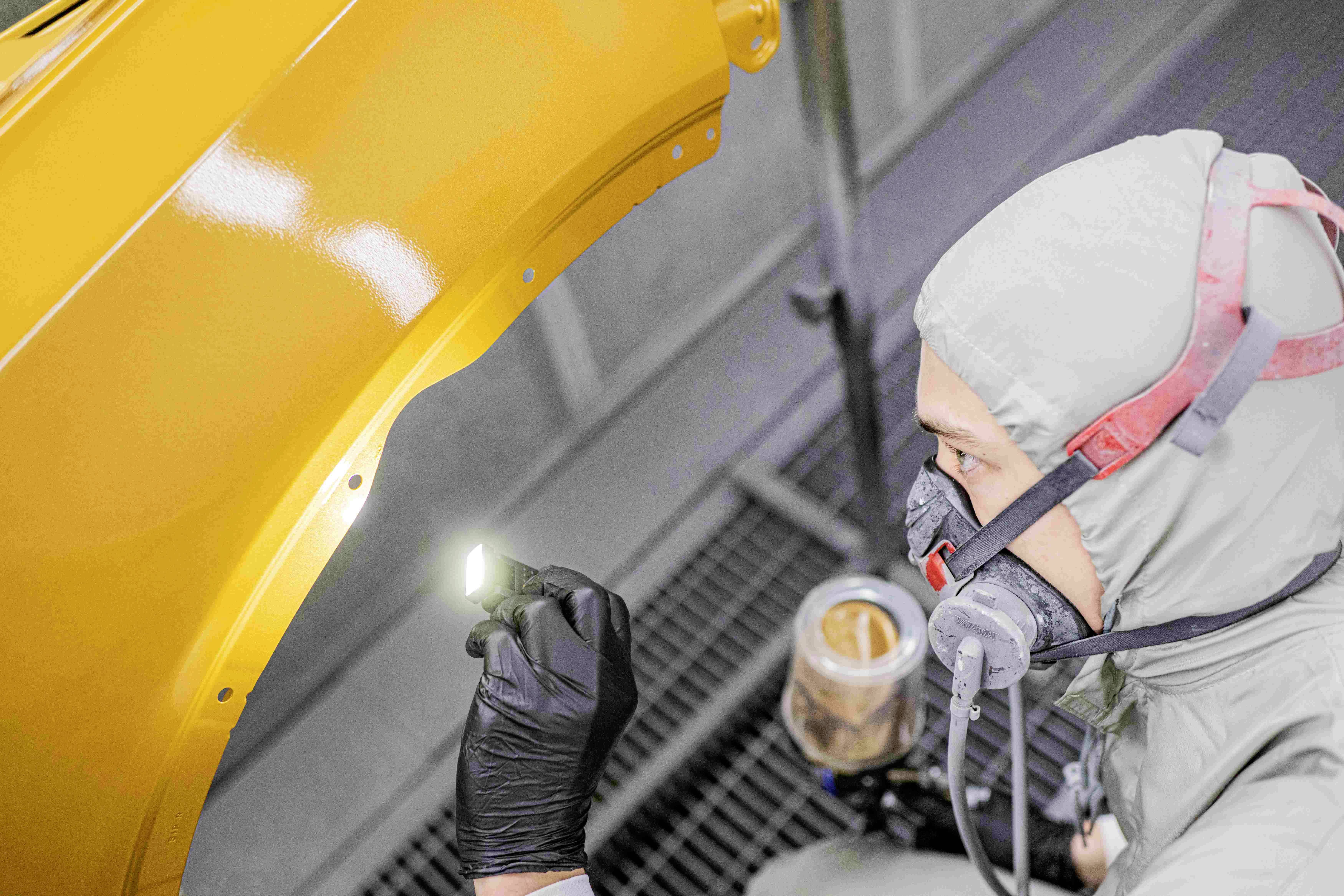 Eine Person in Schutzkleidung inspiziert sorgfältig eine gelbe Autoteiloberfläche in einer Werkstattumgebung, beleuchtet von einer kleinen Lampe.