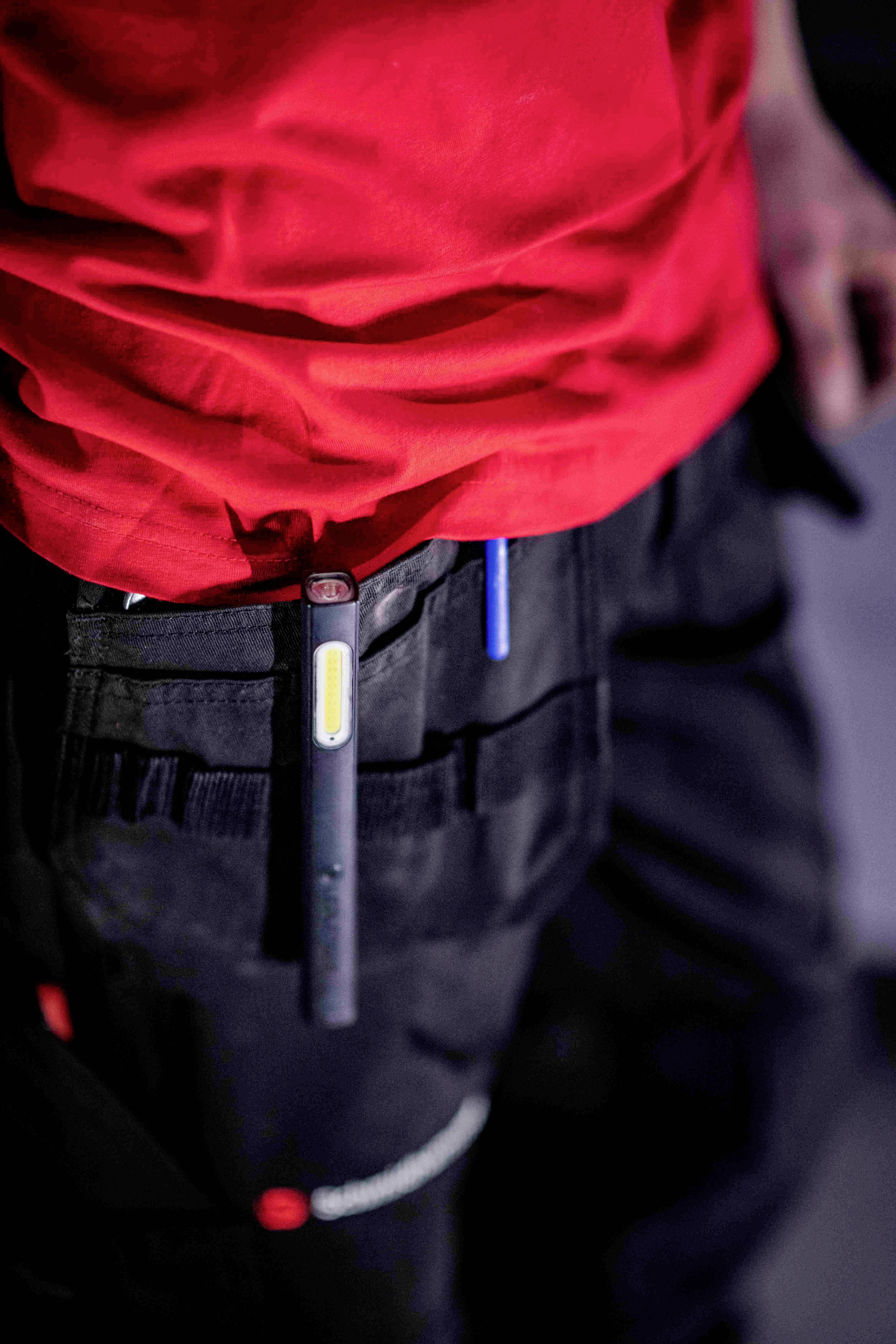 Eine Person trägt eine rote Arbeitsjacke und eine schwarze Hose mit Taschen, in denen zwei Stifte stecken.
