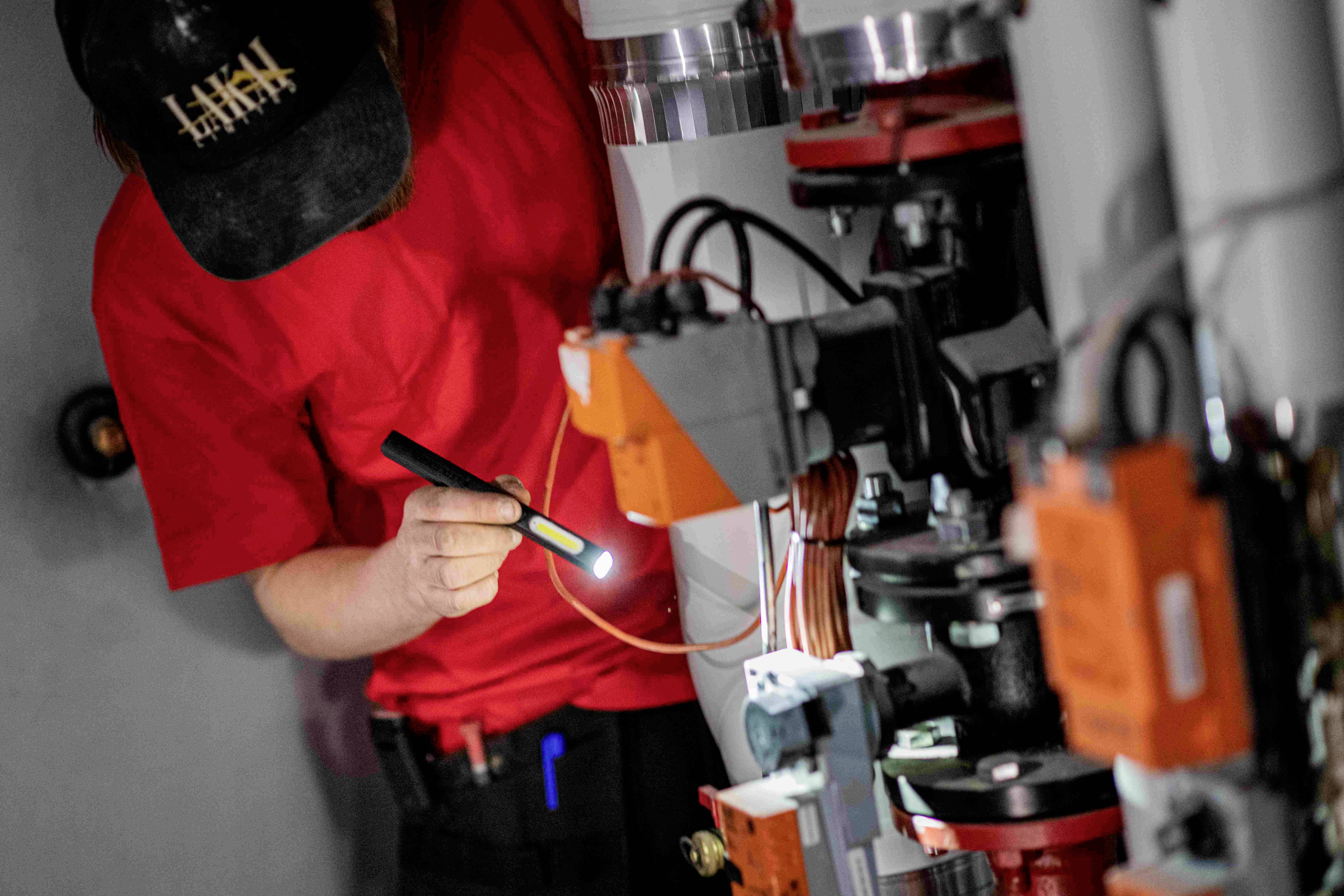 Eine Person in rotem Shirt und Mütze arbeitet mit einer Taschenlampe an einer komplexen Maschine, umgeben von Kabeln und Mechanik.