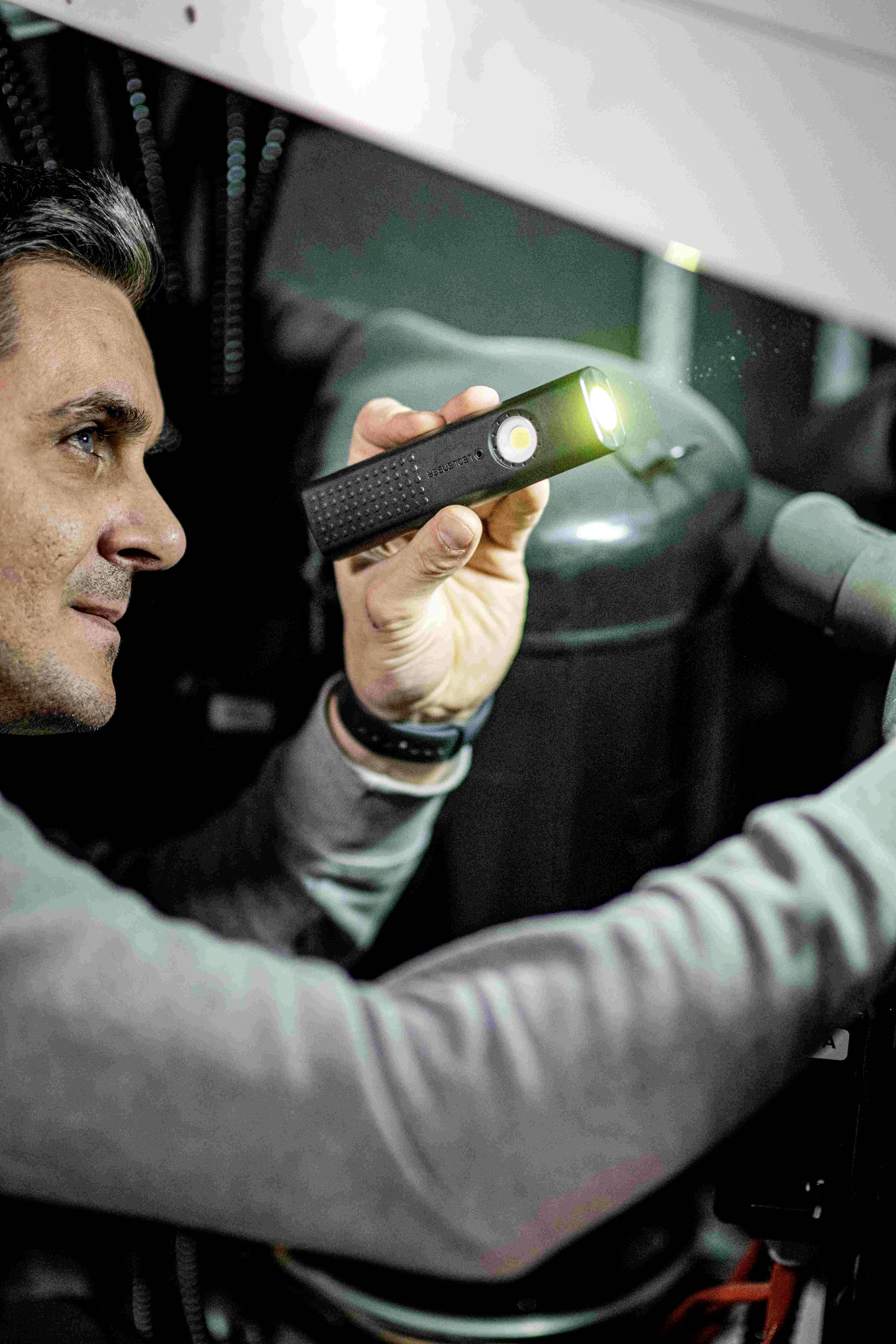 Eine Person inspiziert mit einer Taschenlampe die technischen Komponenten einer Maschine in einer Werkstattumgebung.