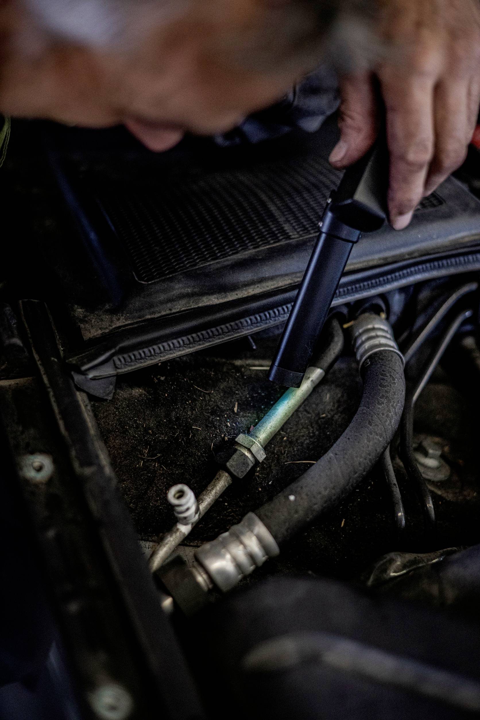 Ein Mechaniker inspiziert einen Autoteil im Motorraum mit einer Taschenlampe. Fokus auf Schläuche und Verbindungen.