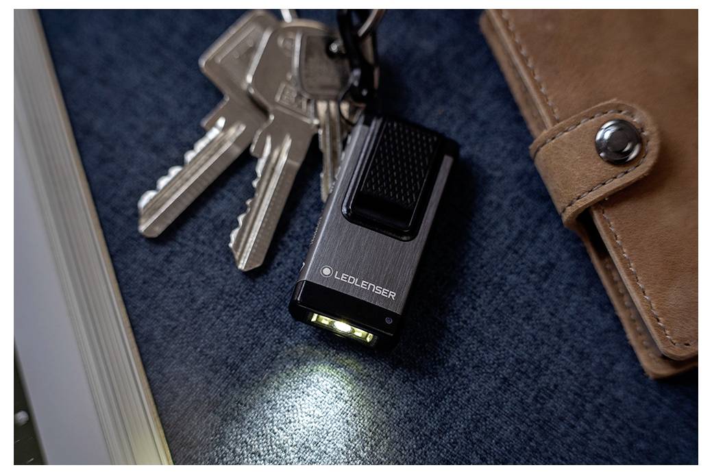 Kleine LEDLENSER Taschenlampe, die auf eine blaue Oberfläche neben Schlüsseln und einer braunen Lederbrieftasche leuchtet.