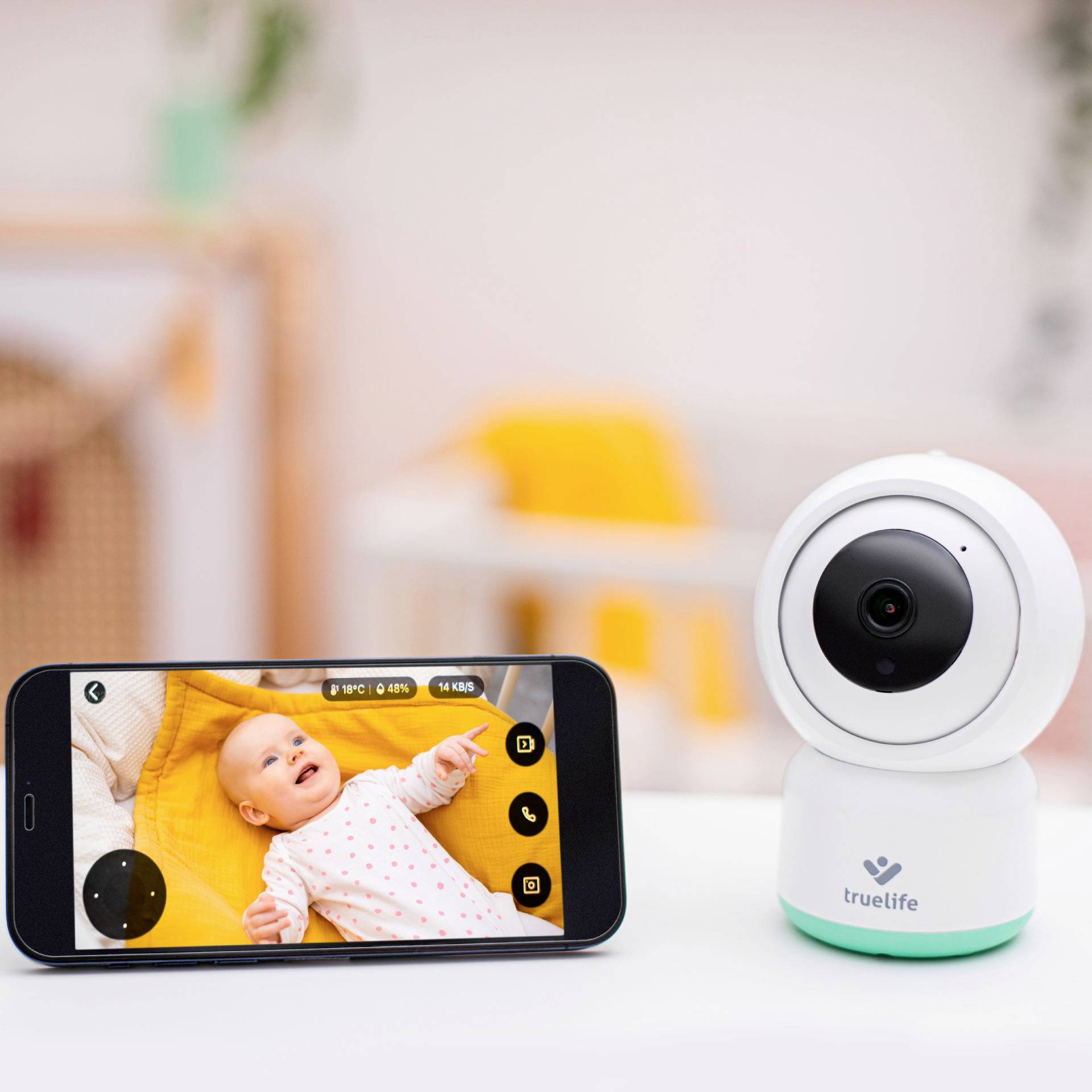 Ein Smartphone zeigt ein Baby auf dem Bildschirm, aufgenommen von einer Baby-Überwachungskamera im Vordergrund.