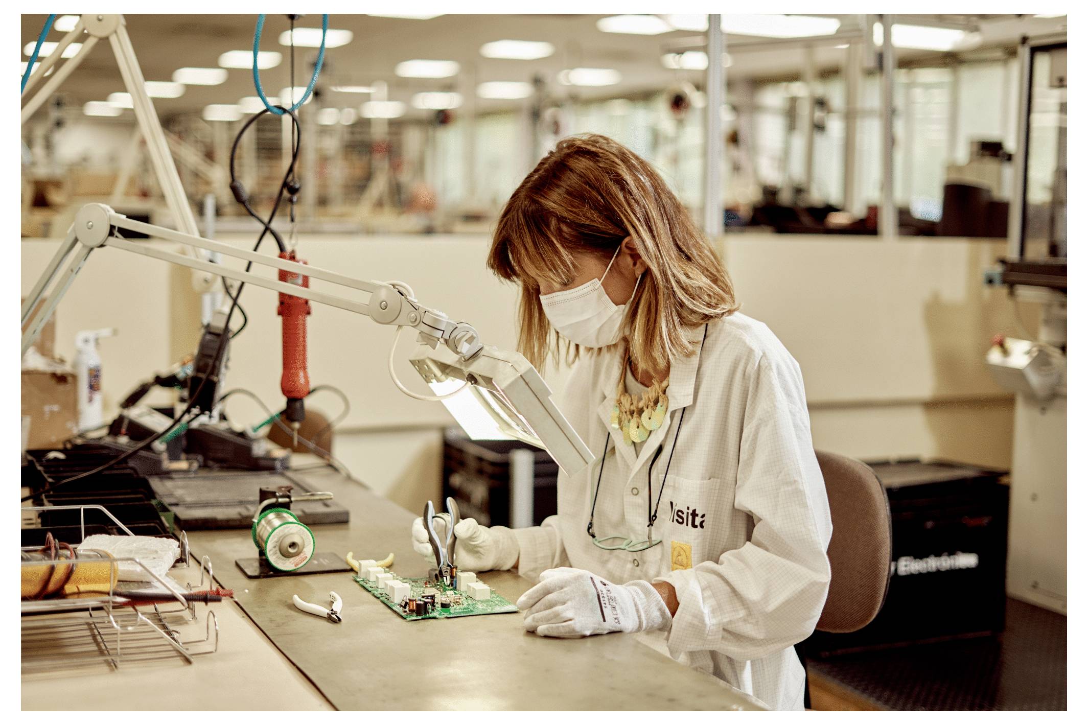 Person wearing a mask and lab coat working on electronic components at a workstation in a manufacturing facility.