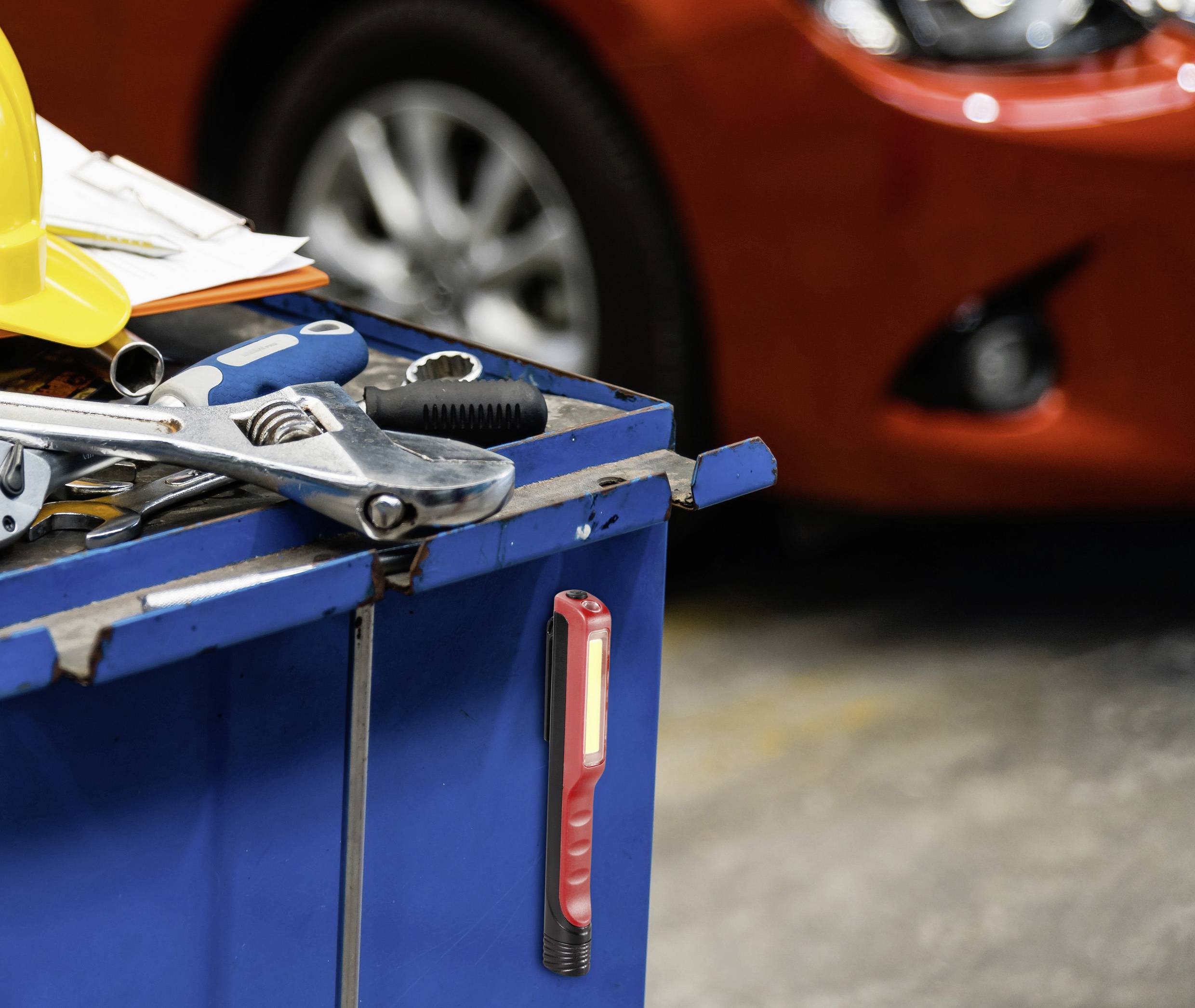 Eine blaue Werkbank mit Werkzeugen, darunter Schraubenschlüssel und ein gelber Helm. Im Hintergrund ein rotes Auto in einer Werkstatt.