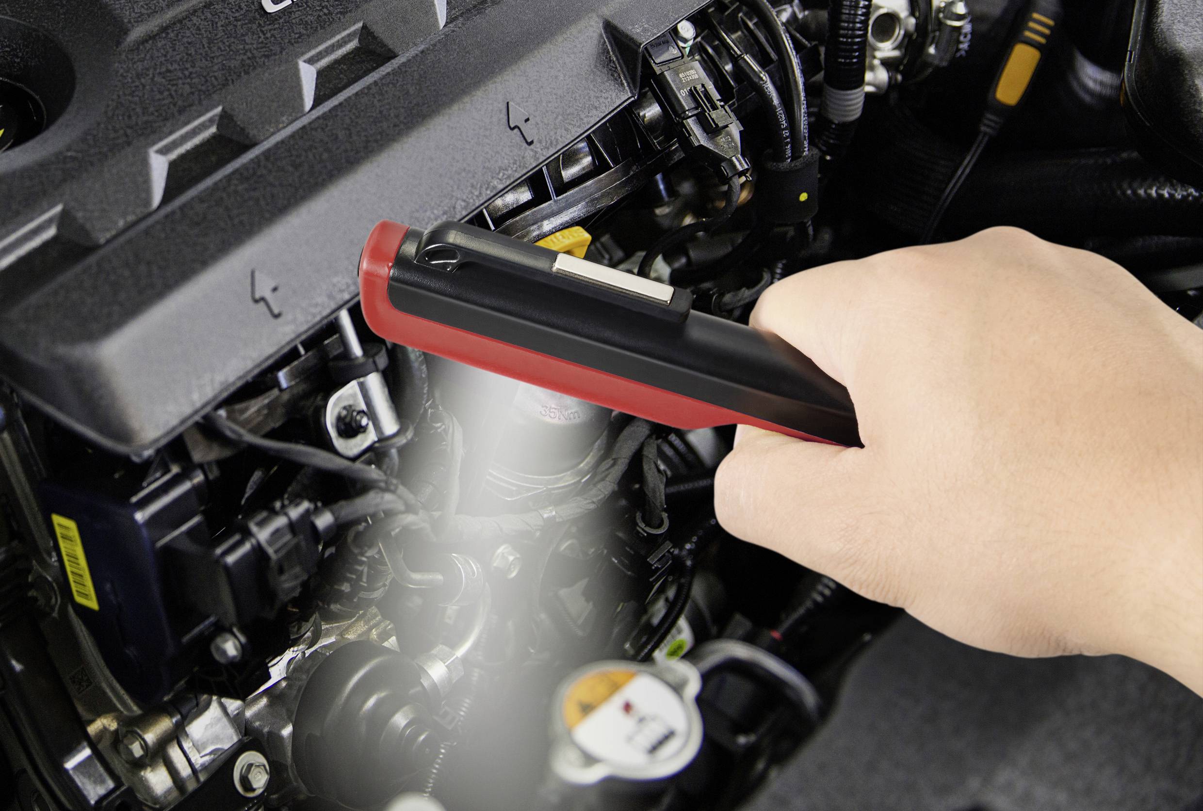 Eine Hand benutzt ein Diagnosetool, um den Motor eines Autos zu prüfen. Das Tool hat ein rotes Gehäuse und wird nah an den Motor gehalten.