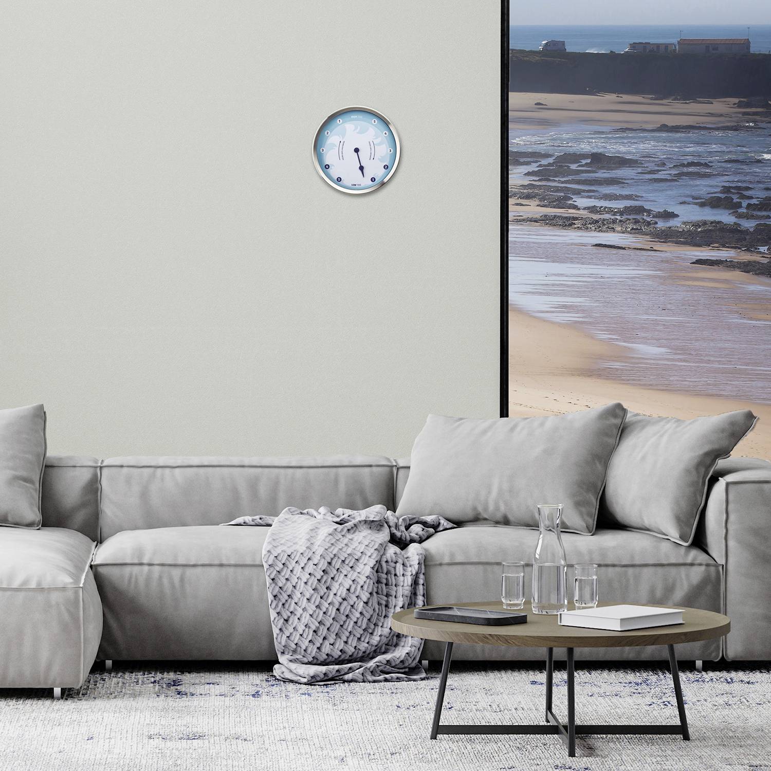 Wohnzimmer mit grauem Sofa, rundem Holztisch, zwei Gläsern und einem Buch, Uhr an der Wand, Fenster mit Blick auf Strand und Meer.