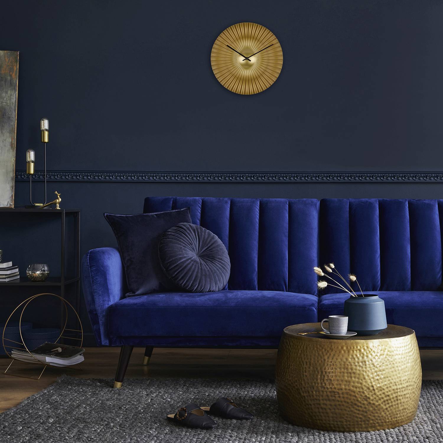 Ein stilvolles Wohnzimmer mit einem blauen Sofa, verzierten Wänden und einem goldenen runden Tisch. An der Wand hängt eine goldene Uhr.