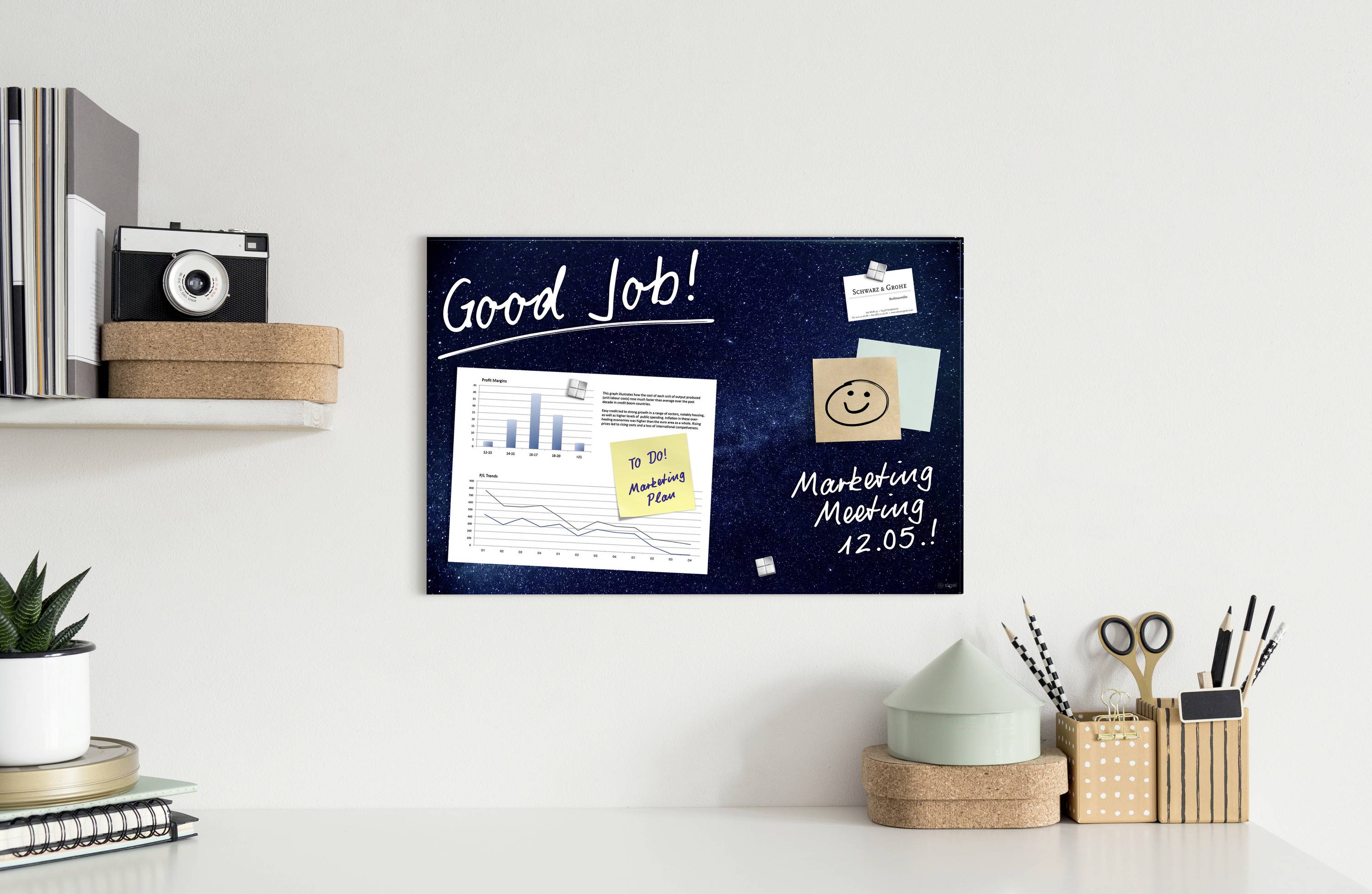 Auf einem Schreibtisch steht ein Regal mit Kamera und Pflanzen. An der Wand hängt eine Pinnwand mit Notizen, Grafiken und dem Text 'Good Job!' und 'Marketing Meeting 12.05.'.