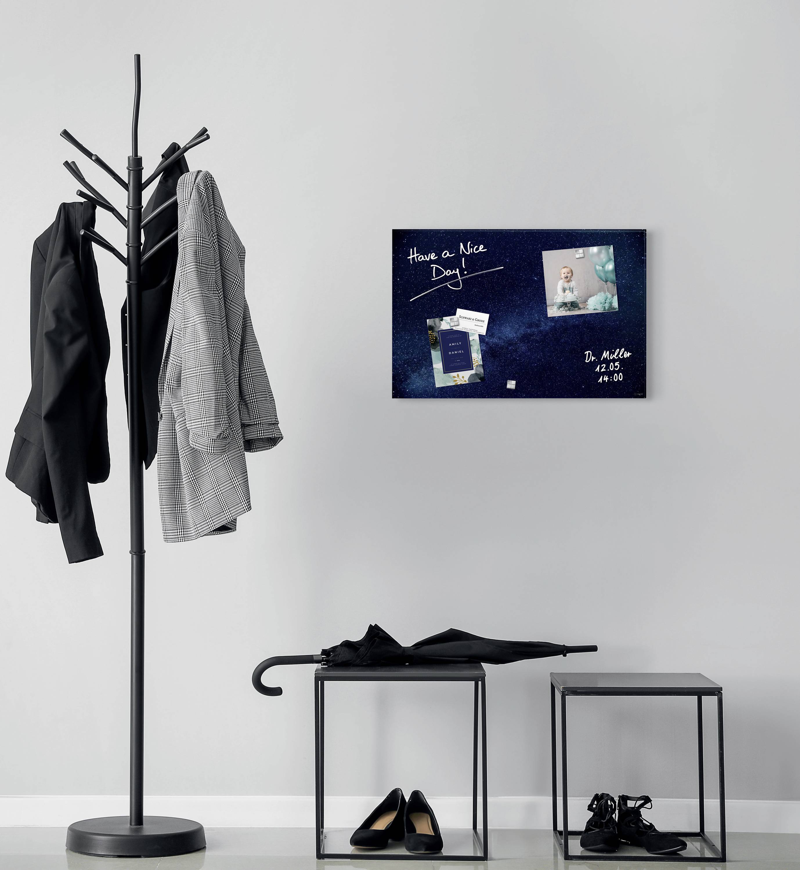 Garderobenständer mit Jacken und eine Pinnwand an der Wand mit 'Have a Nice Day' und einem Foto, daneben schwarze Hocker.