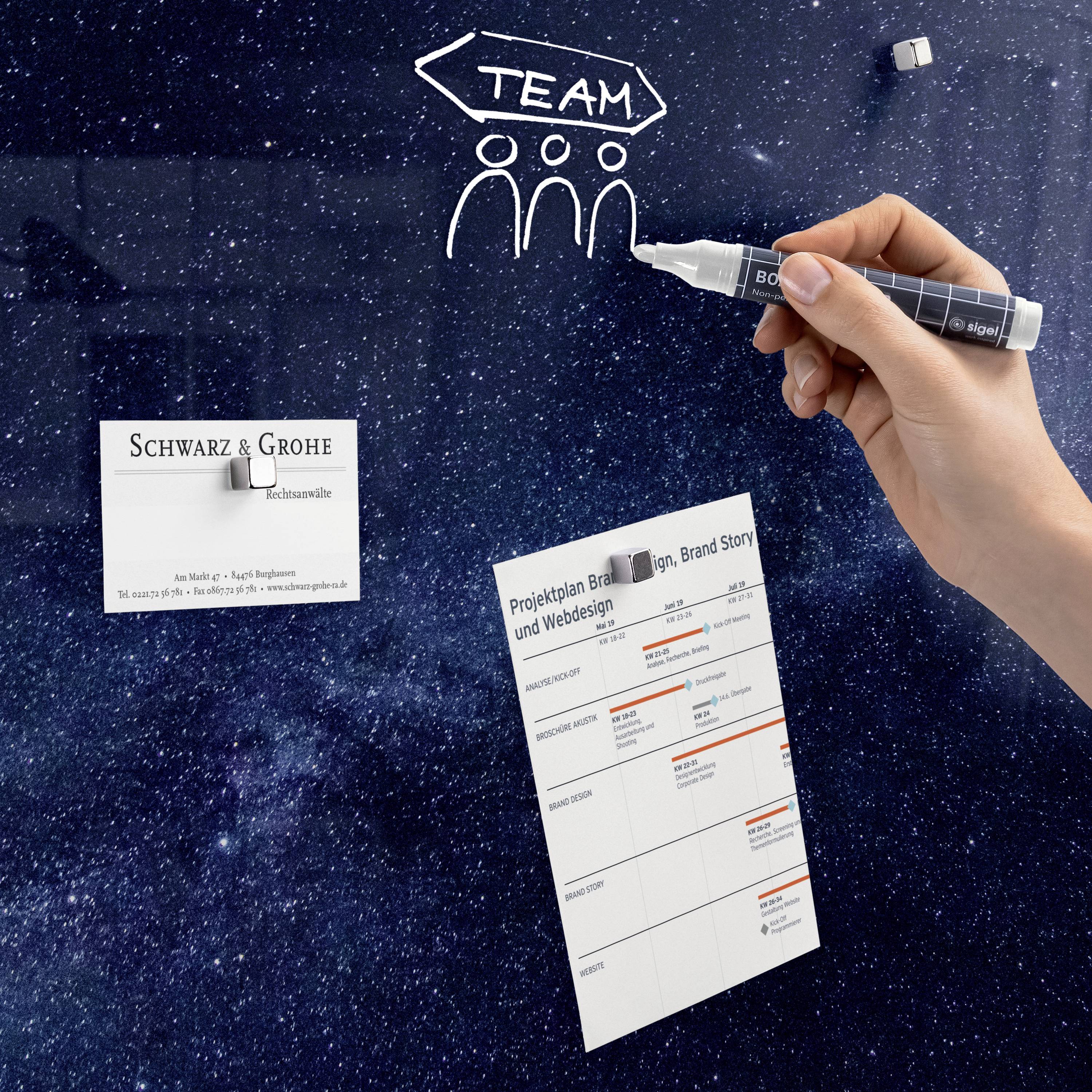 Eine Hand zeichnet mit einem Stift auf einer Tafel 'Team' über drei stilisierte Personen. Unten sind eine Visitenkarte und ein Projektplan sichtbar.