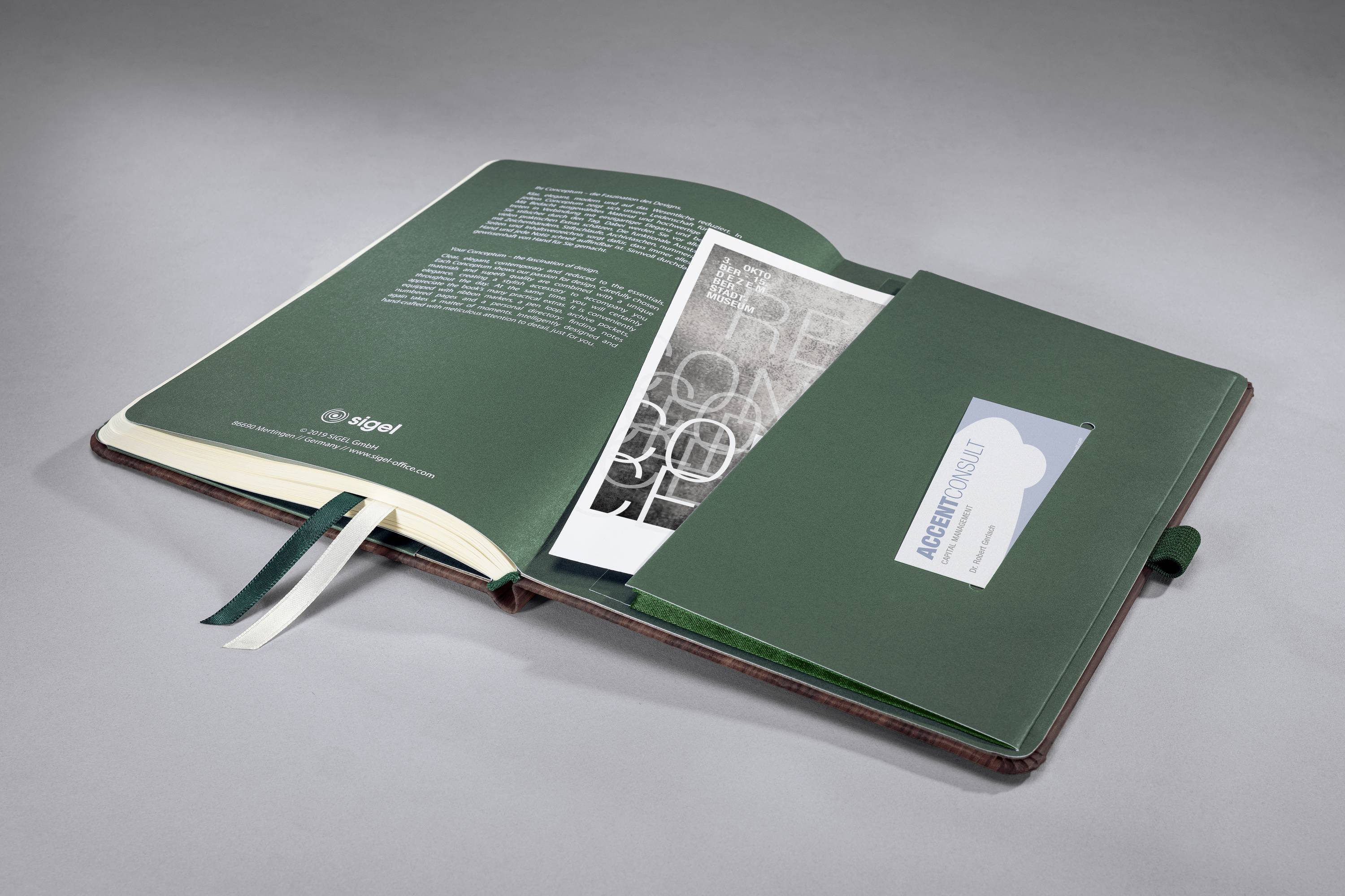 Offenes Notizbuch mit grünem Einband, zeigt eine Textseite und eine Einstecktasche rechts, die ein Dokument und eine Visitenkarte enthält.