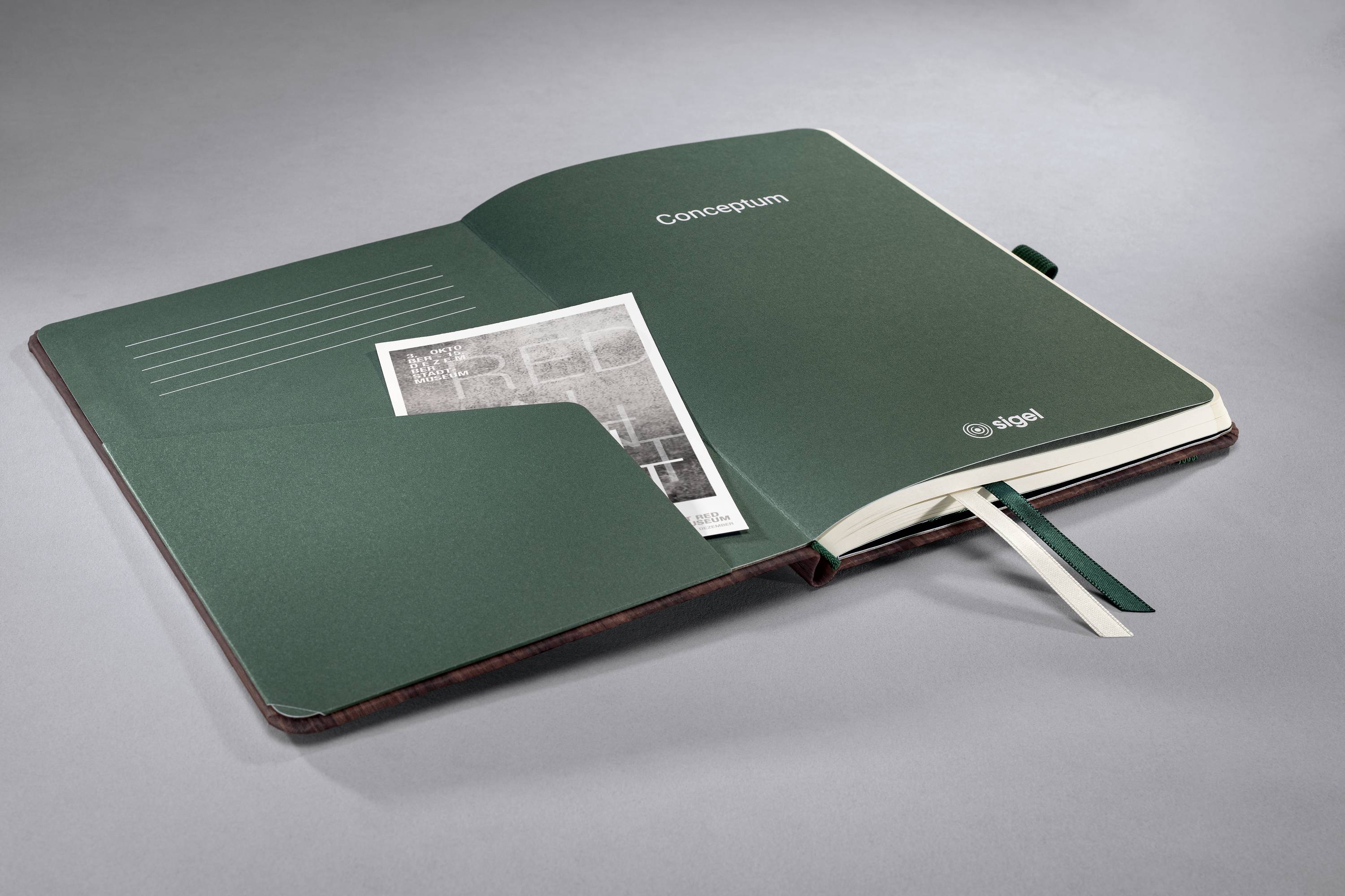 Ein aufgeschlagenes Notizbuch mit grünem Einband. Innen steckt eine schwarz-weiße Zeitungsseite in einer Tasche des Einbands.