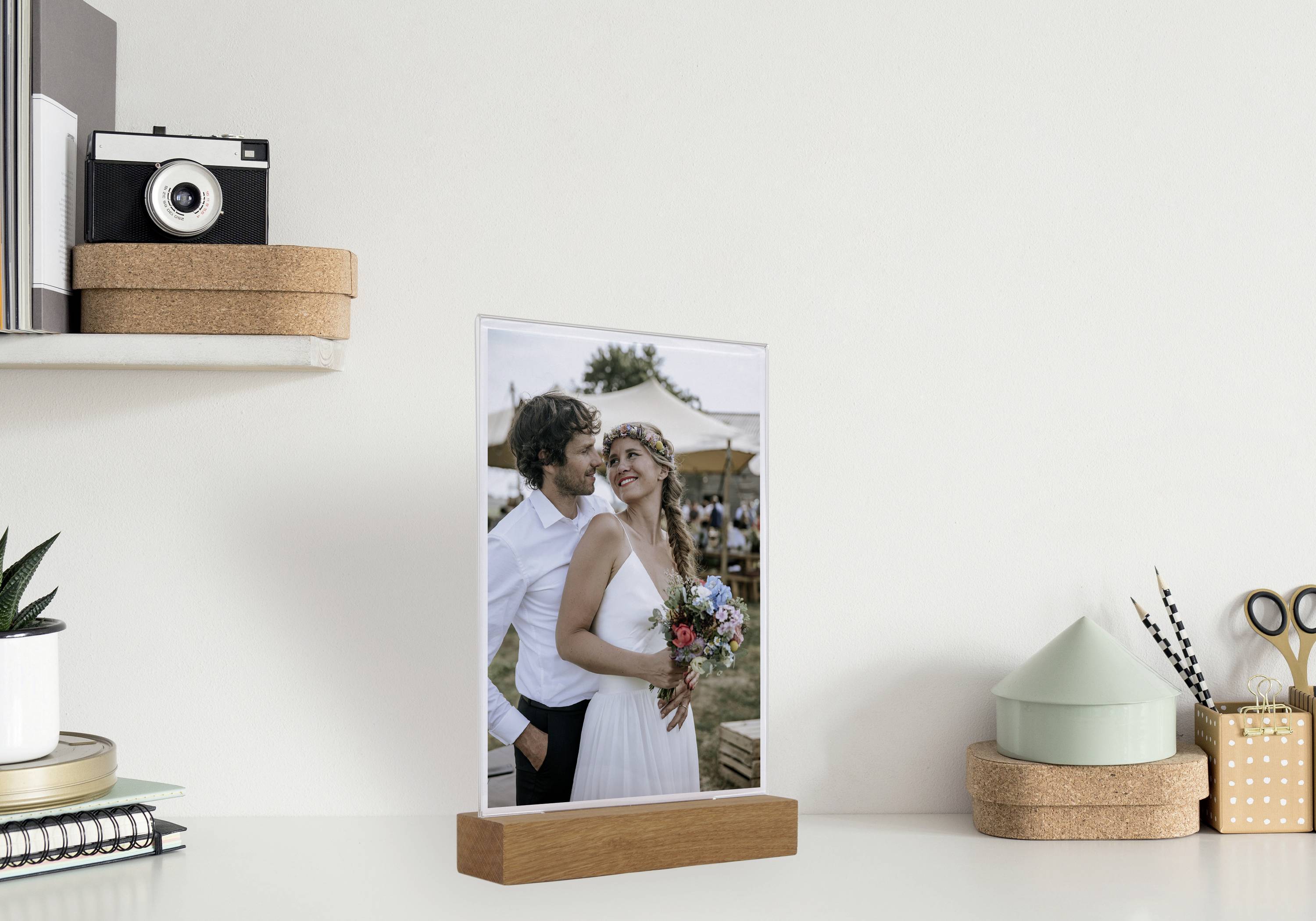 Ein Hochzeitsfoto auf einem Holzrahmen zeigt ein lächelndes Paar, wobei der Mann die Frau umarmt. Im Hintergrund sind Bäume und Zelte sichtbar.