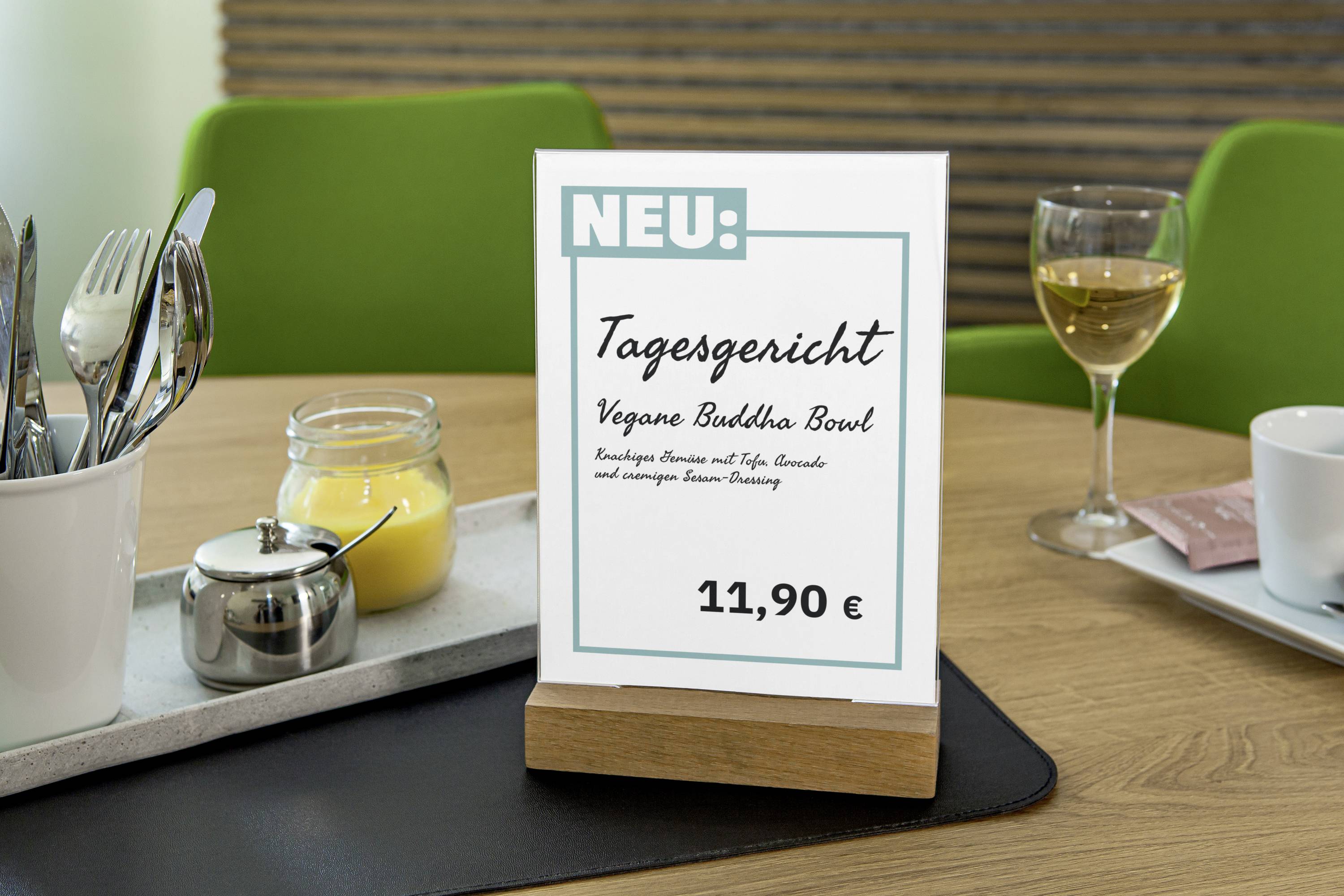 Tischaufsteller im Restaurant zeigt Tagesgericht: 'Vegane Buddha Bowl' für 11,90 EUR. Im Hintergrund Wein und Besteck.