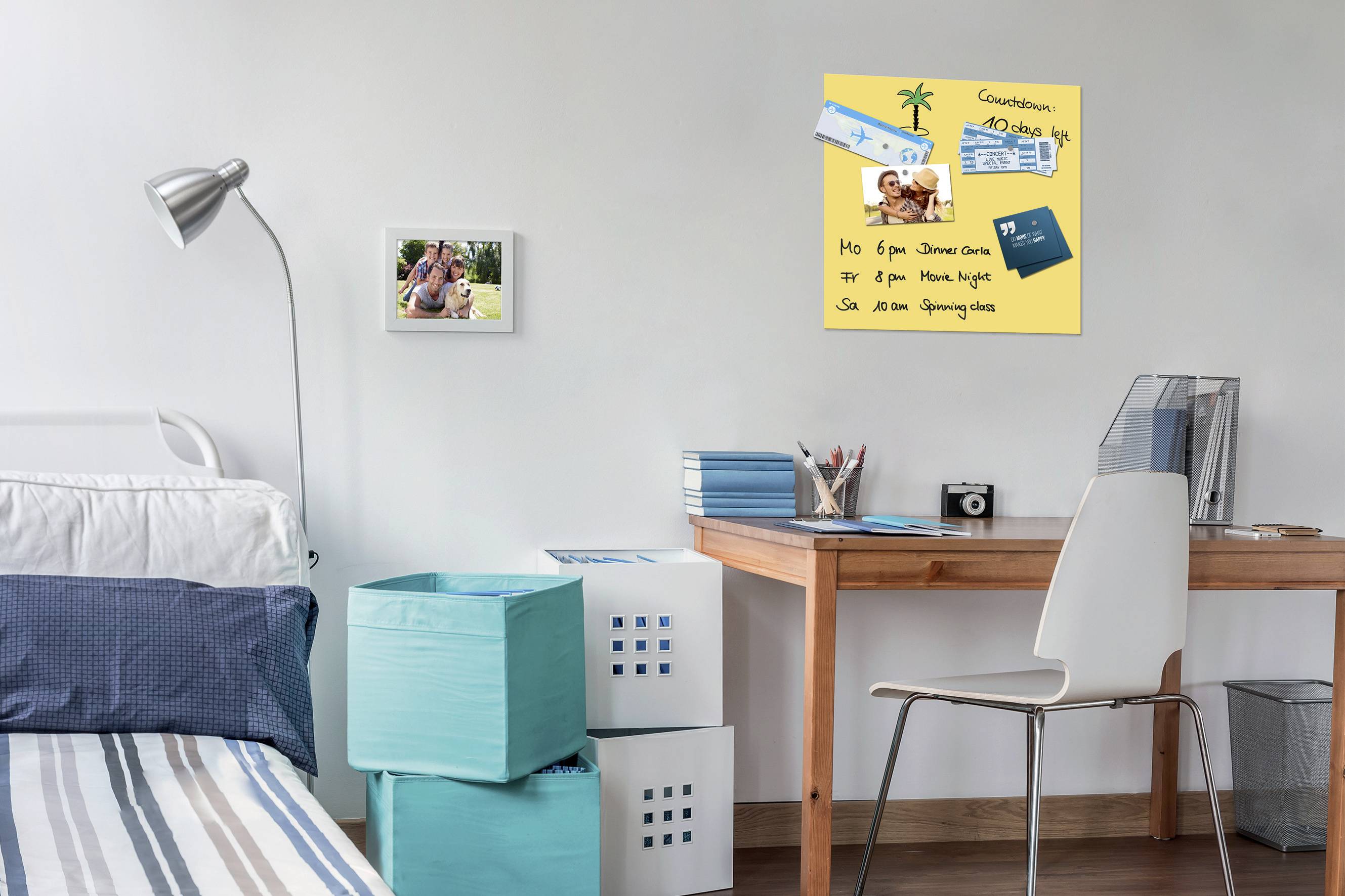 Ein Schreibtisch mit Stuhl steht in einem Schlafzimmer. An der Wand hängt eine Notiztafel mit Fotos, Notizen und einer Eintrittskarte.