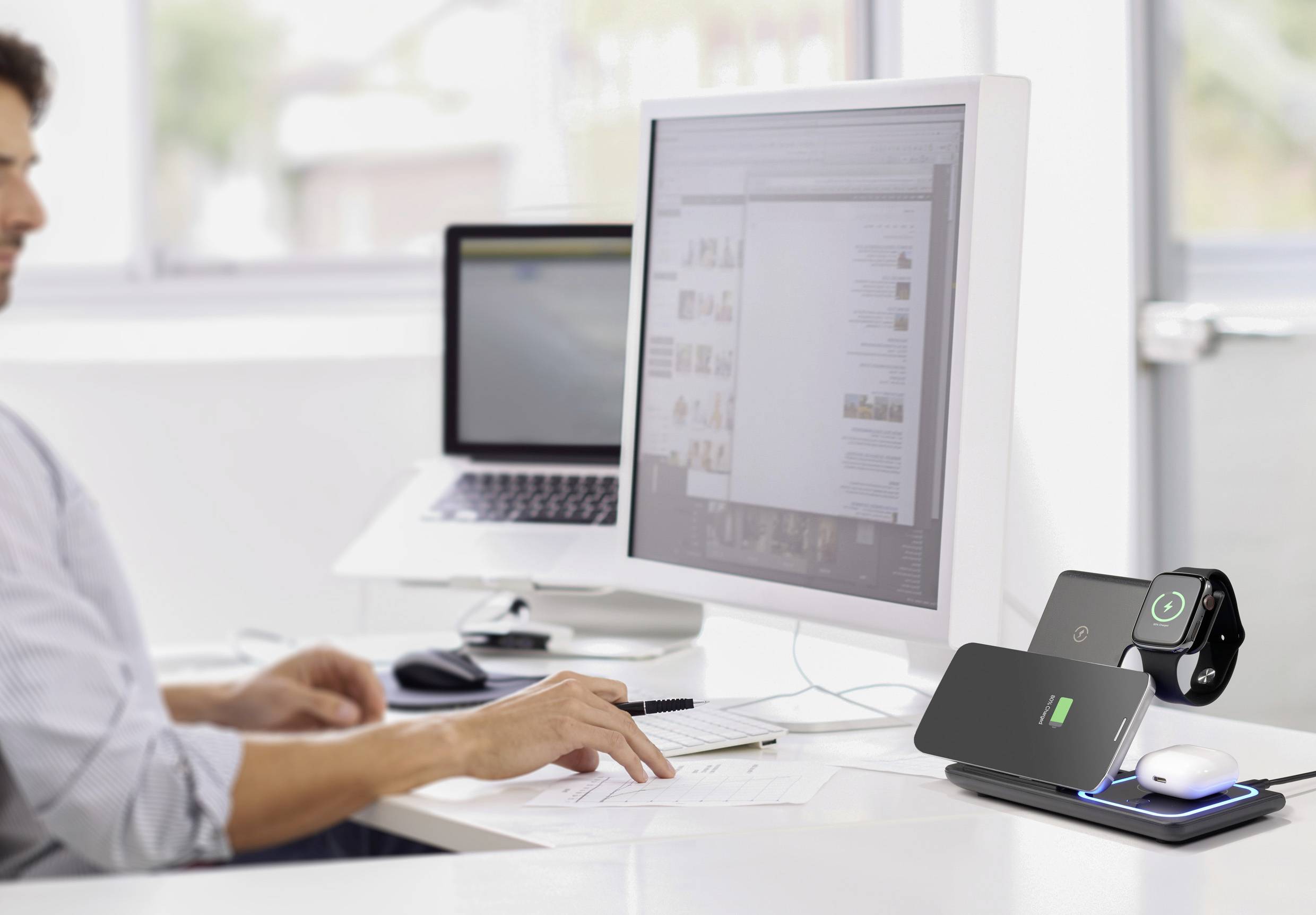 Ein Mann arbeitet am Computer in einem modernen Büro. Im Vordergrund ist eine Ladestation mit einem Smartphone und einer Smartwatch.