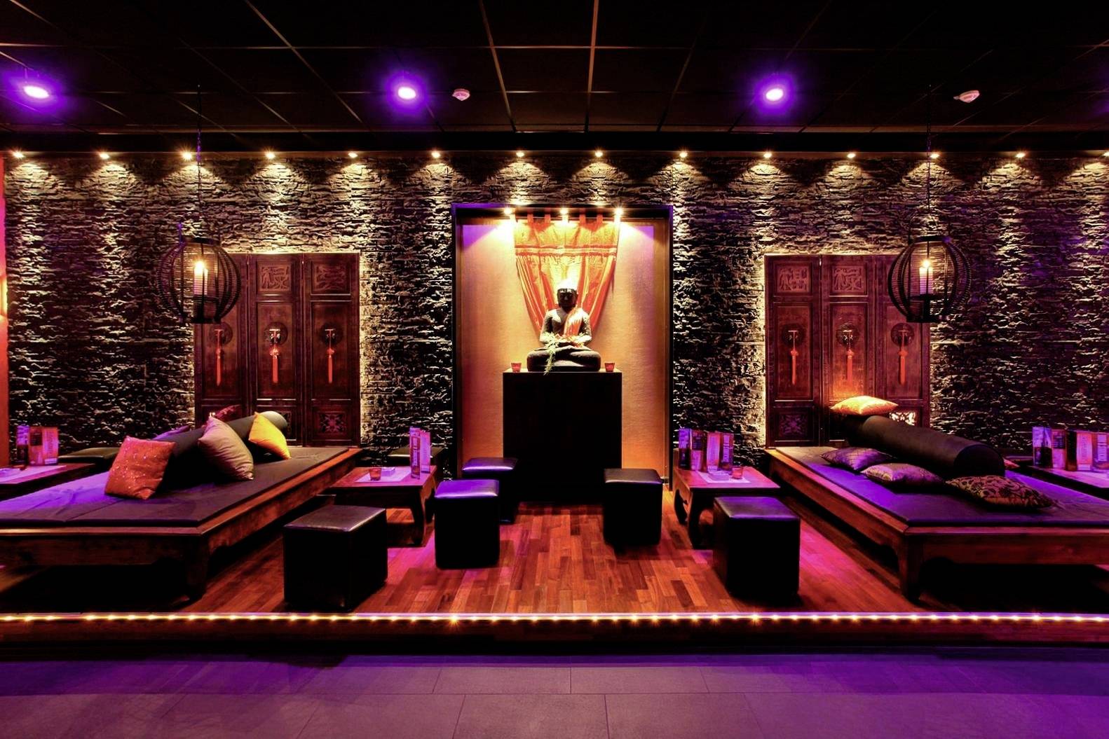 Ein stilvoll beleuchteter Loungebereich mit niedrigen Sitzgelegenheiten und dekorativen Kissen, umgeben von steinernen Wänden und warmem Licht.