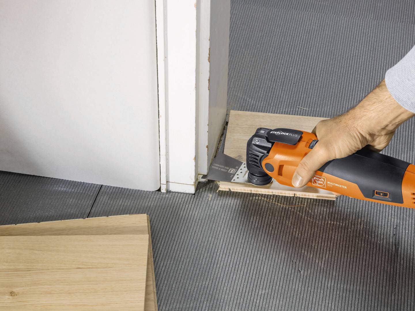Eine Person schneidet mit einem Multifunktionswerkzeug eine Holzplatte am Boden, um sie passend an den Türrahmen anzupassen.