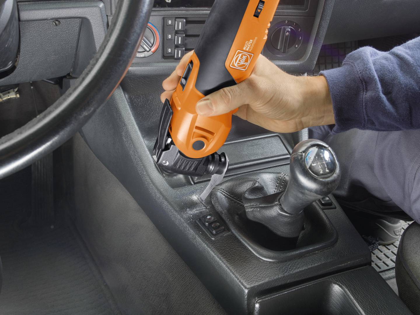 Eine Person reinigt die Mittelkonsole eines Autos mit einem orangefarbenen, akkubetriebenen Multitool.