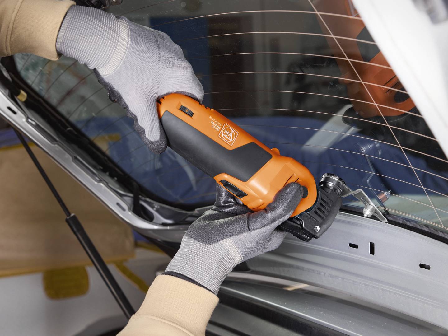Eine Person verwendet ein orangefarbenes Elektrowerkzeug, um an der Heckscheibe eines Autos zu arbeiten. Sie trägt graue Handschuhe.