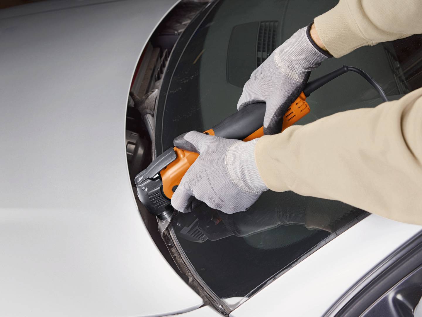 Eine Person schneidet mit einem elektrischen Werkzeug die Windschutzscheibe eines Autos heraus. Hände tragen Schutzhandschuhe.