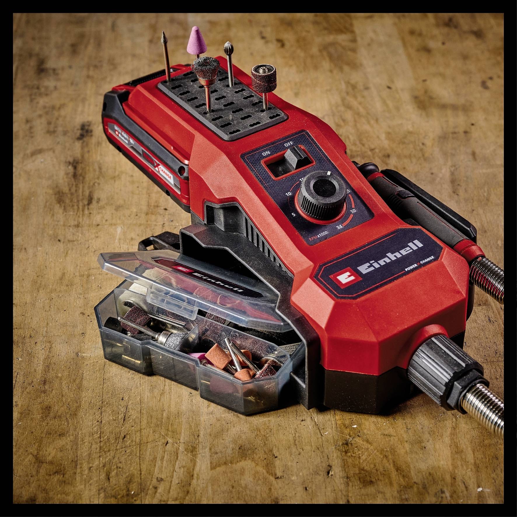Ein rotes, tragbares Multitool mit verschiedenen Bohr- und Schleifaufsätzen auf einem Holztisch. Zubehör ist in einer transparenten Box.