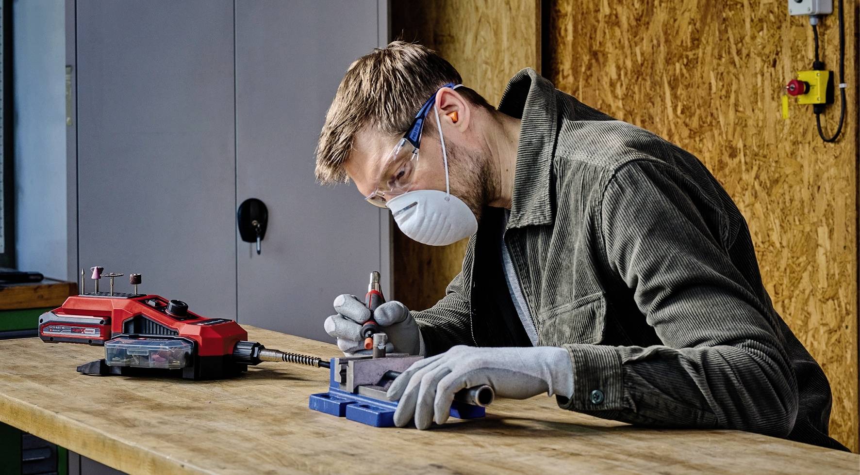 Ein Mann arbeitet konzentriert an einem Tisch, trägt eine Schutzmaske und Handschuhe und benutzt ein Werkzeug für präzise Handarbeit.