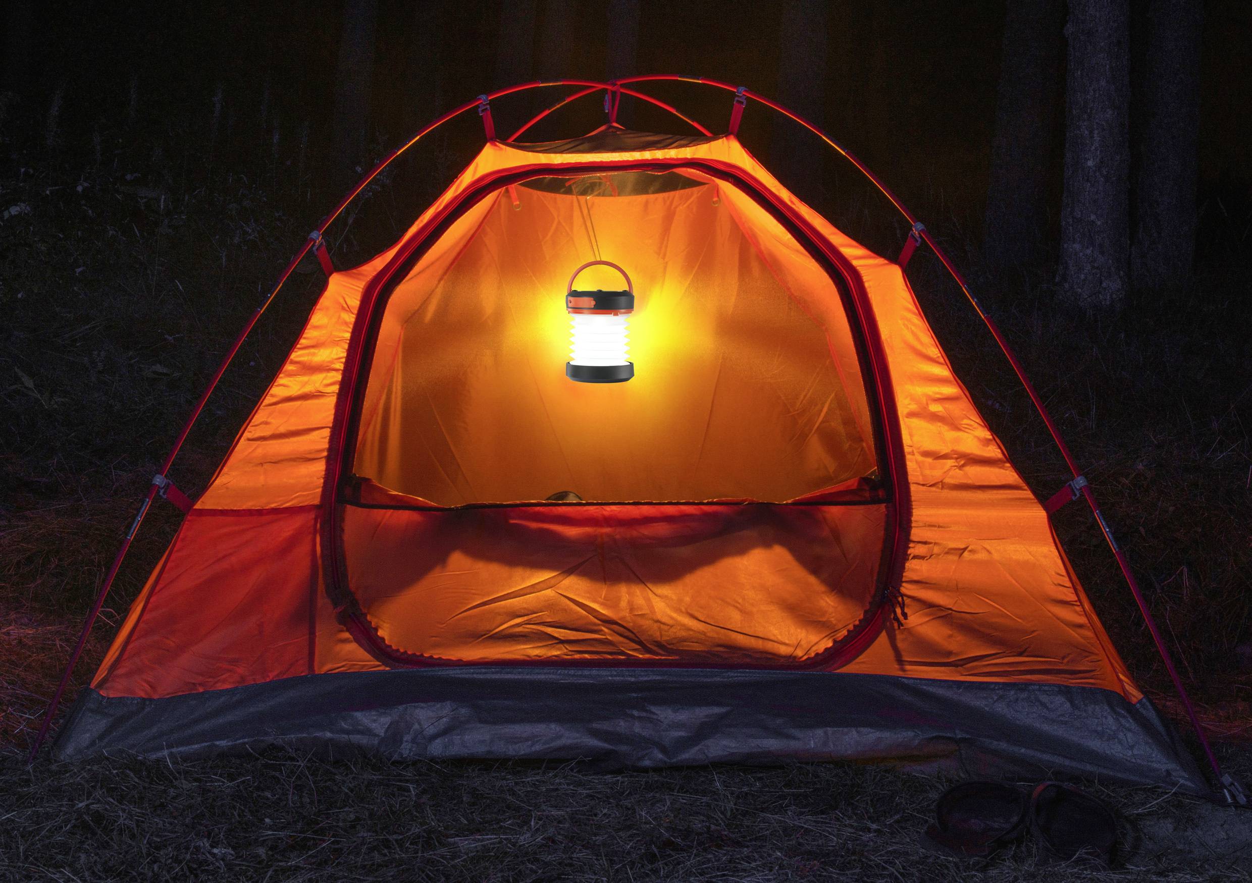 Ein orangefarbenes Zelt steht im Dunkeln, beleuchtet von einer hellen Laterne im Inneren, die den Eingang erhellt.