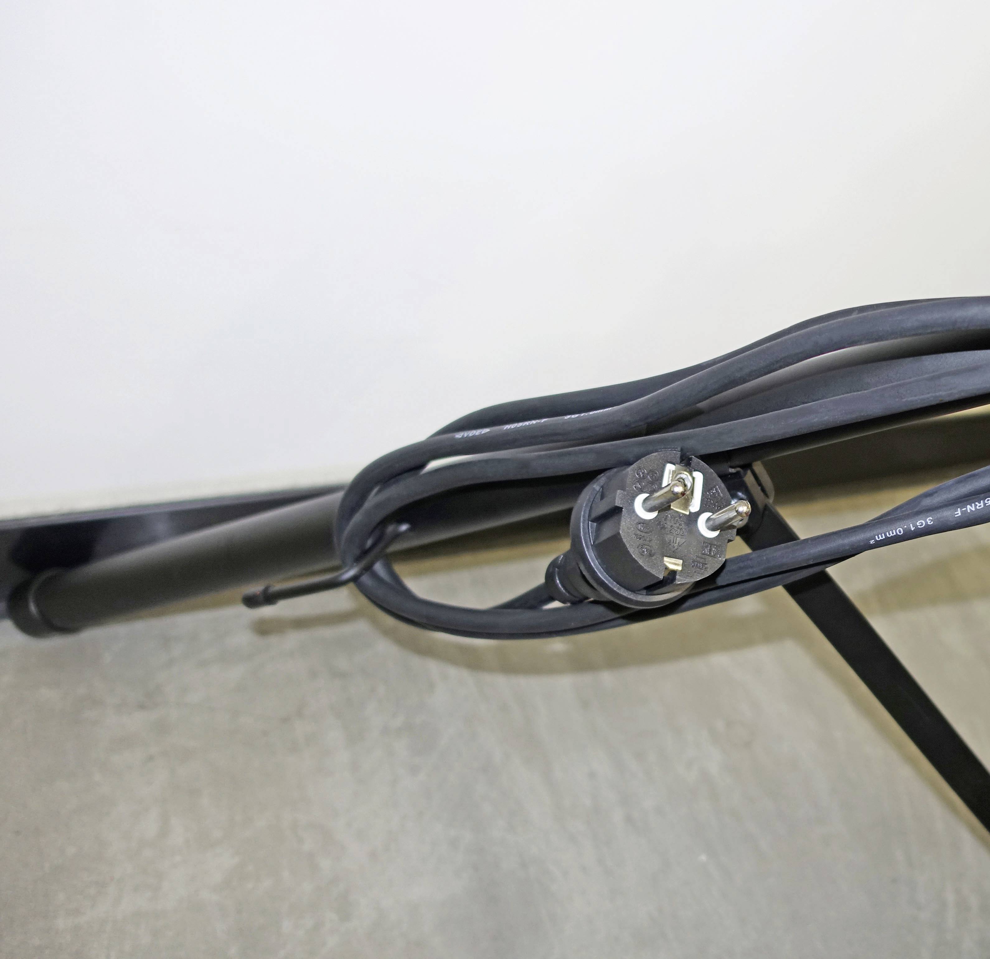 Ein schwarzes Ladekabel mit einem dreipoligen Stecker liegt auf einem grauen Boden. Es ist teilweise aufgewickelt und führt zu einer Ladestation.