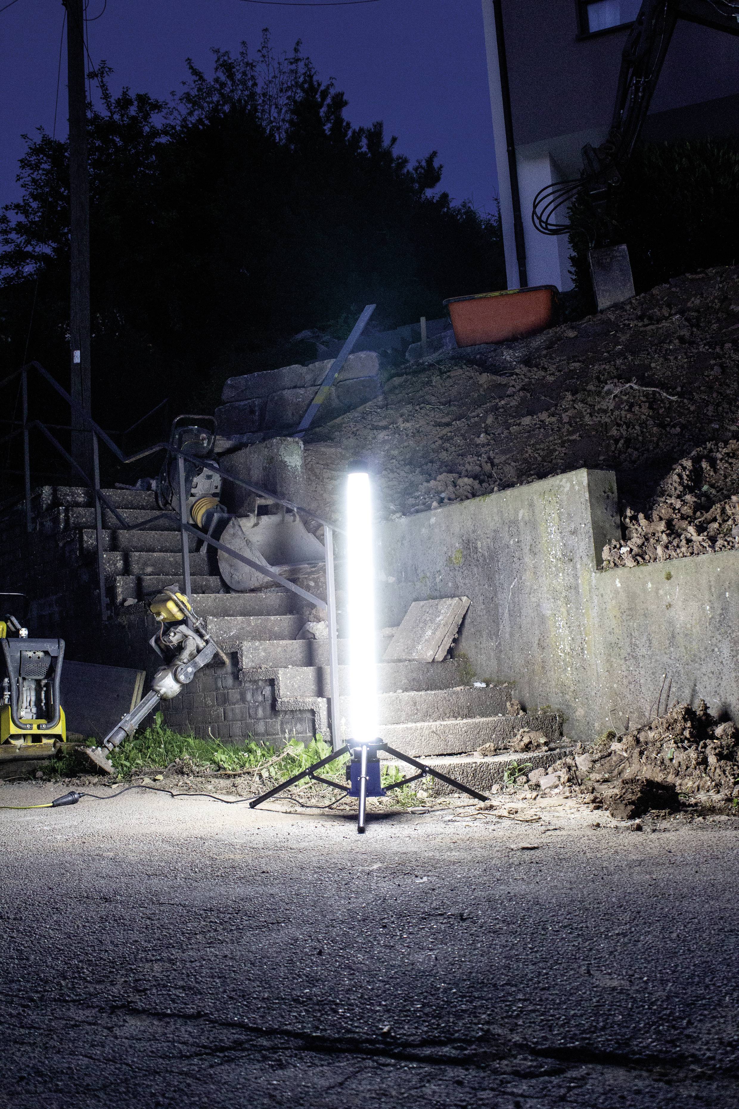 Eine Baustelle bei Nacht mit einer hellen Arbeitsleuchte, die eine steinige Treppe und umgebende Erde beleuchtet. Werkzeuge sind verstreut.