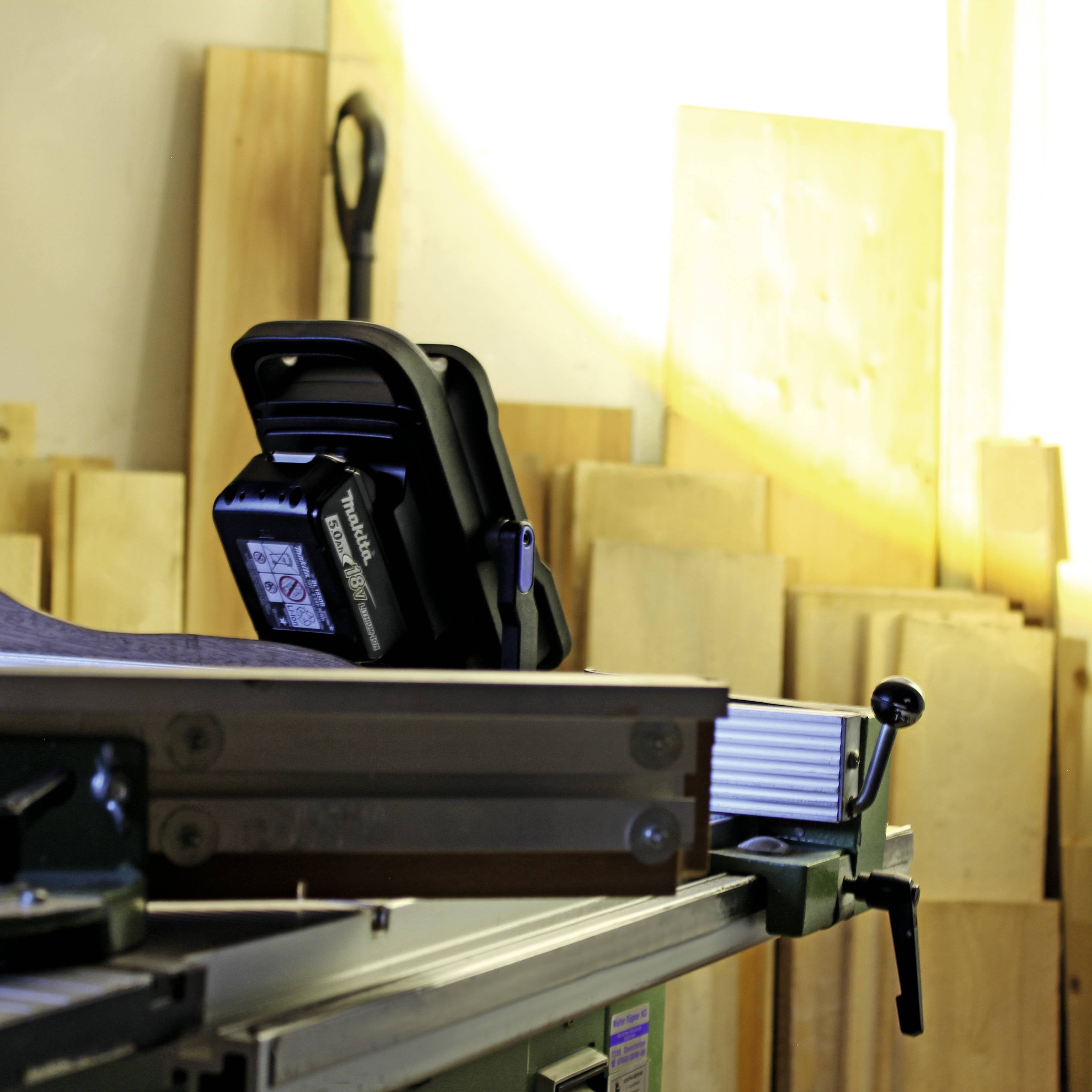 In einer Werkstatt steht eine Tischkreissäge. Im Hintergrund sind Holzplatten und Werkzeug zu sehen. Ein Maßband liegt auf der Säge.