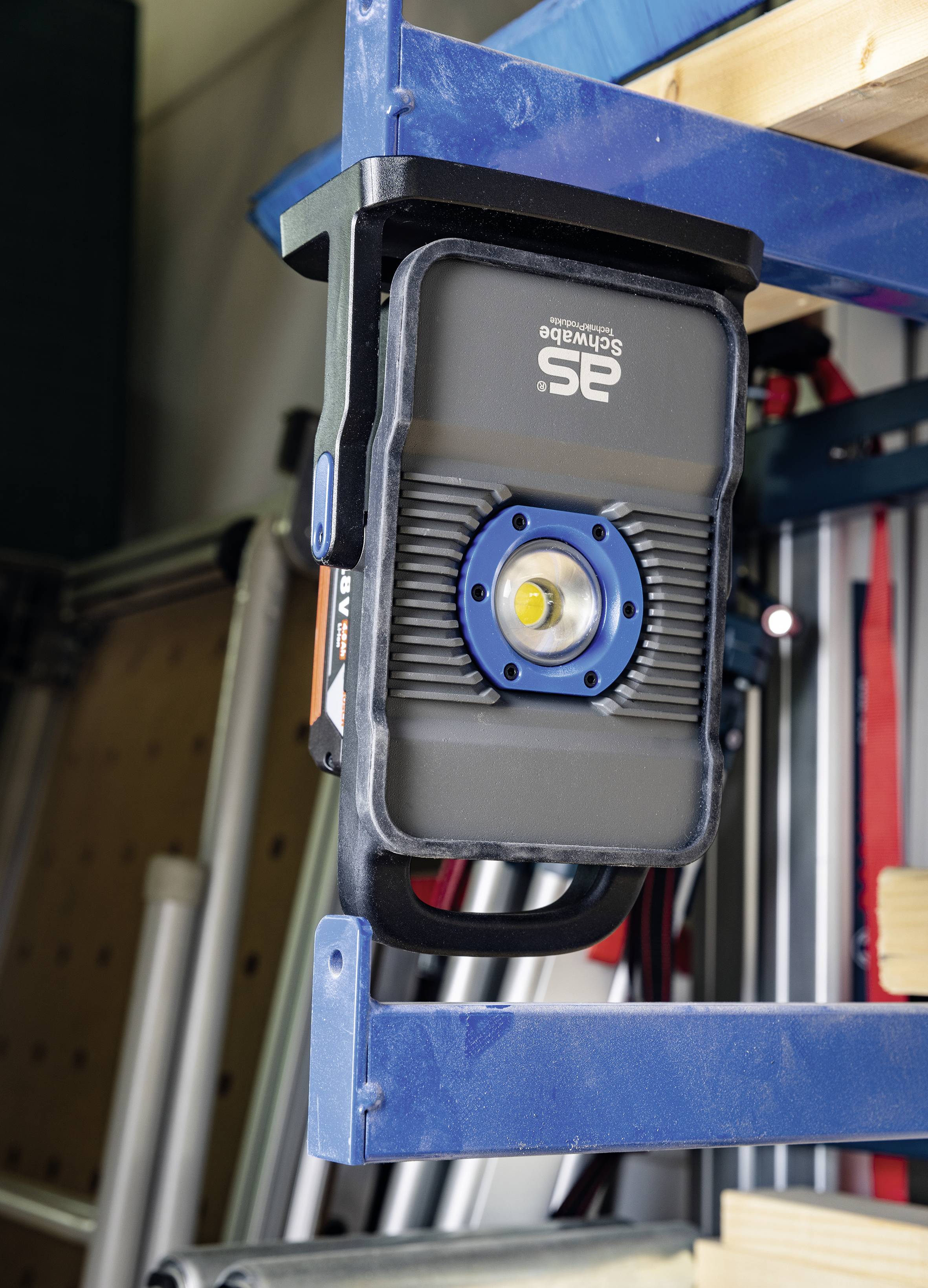 Eine schwarze LED-Arbeitsleuchte ist an einem blauen Regal befestigt. Im Hintergrund sind Metallleitern und Holzlatten sichtbar.
