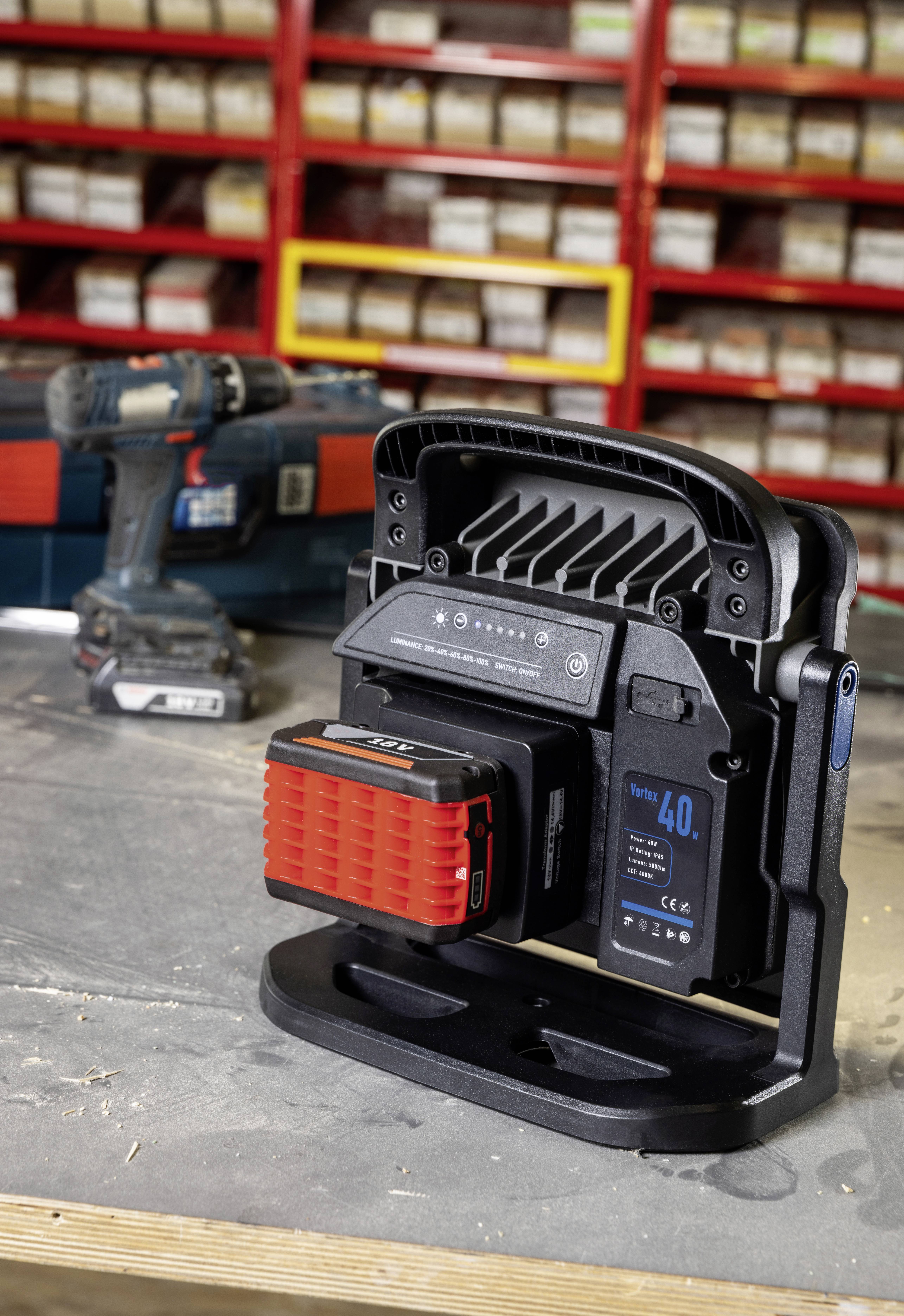 Ein tragbares LED-Arbeitslicht mit aufgesetztem Akku steht auf einer Werkbank. Im Hintergrund sind Regale mit Schachteln und eine Bohrmaschine.