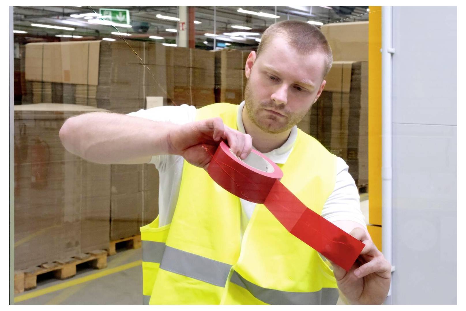 Ein Mann in einer Warnweste klebt rotes Klebeband auf eine Glasfläche in einem Lagerhaus, während im Hintergrund Stapel von Kartons stehen.