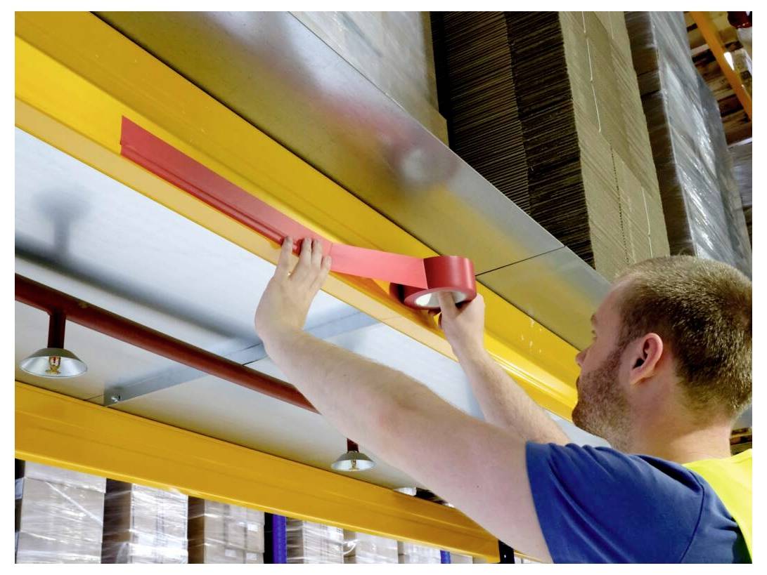 Eine Person in einem Lagerhaus befestigt rotes Klebeband an einem Metallträger unter einem Regal, wobei Stapel von Kartons darüber sichtbar sind.