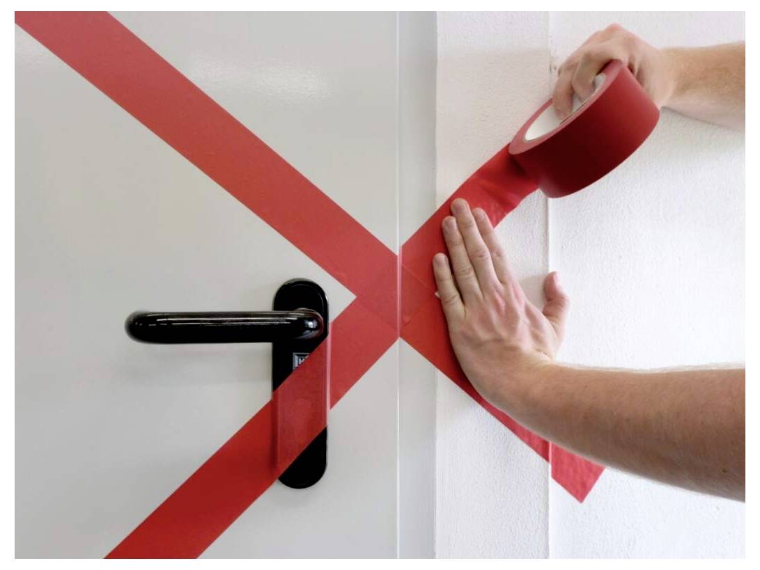 Eine Person, die rotes Klebeband in Form eines 'X' über eine Tür und Wand klebt und dabei den Türgriff abdeckt.