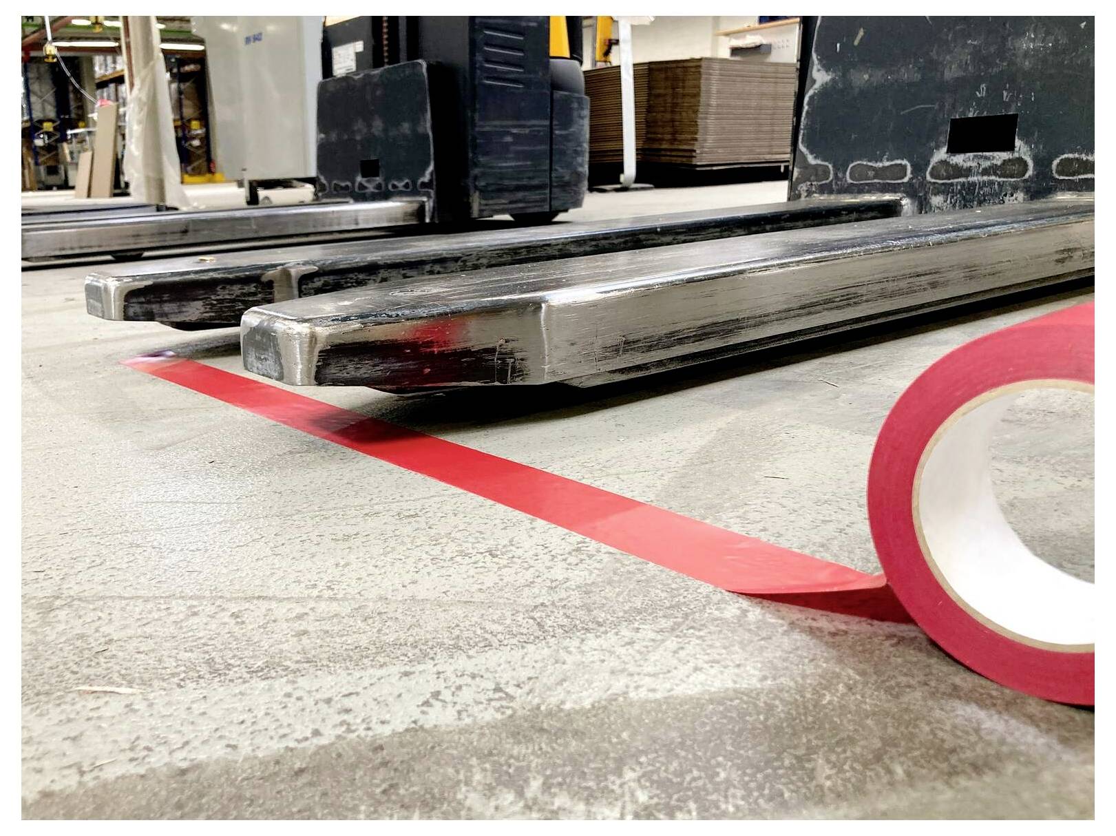 „Nahaufnahme eines Gabelstaplers, dessen Gabeln über rotem Bodenmarkierungsband in einem Lagerhaus ausgerichtet sind, mit Betonung auf präziser Positionierung für die Sicherheit.