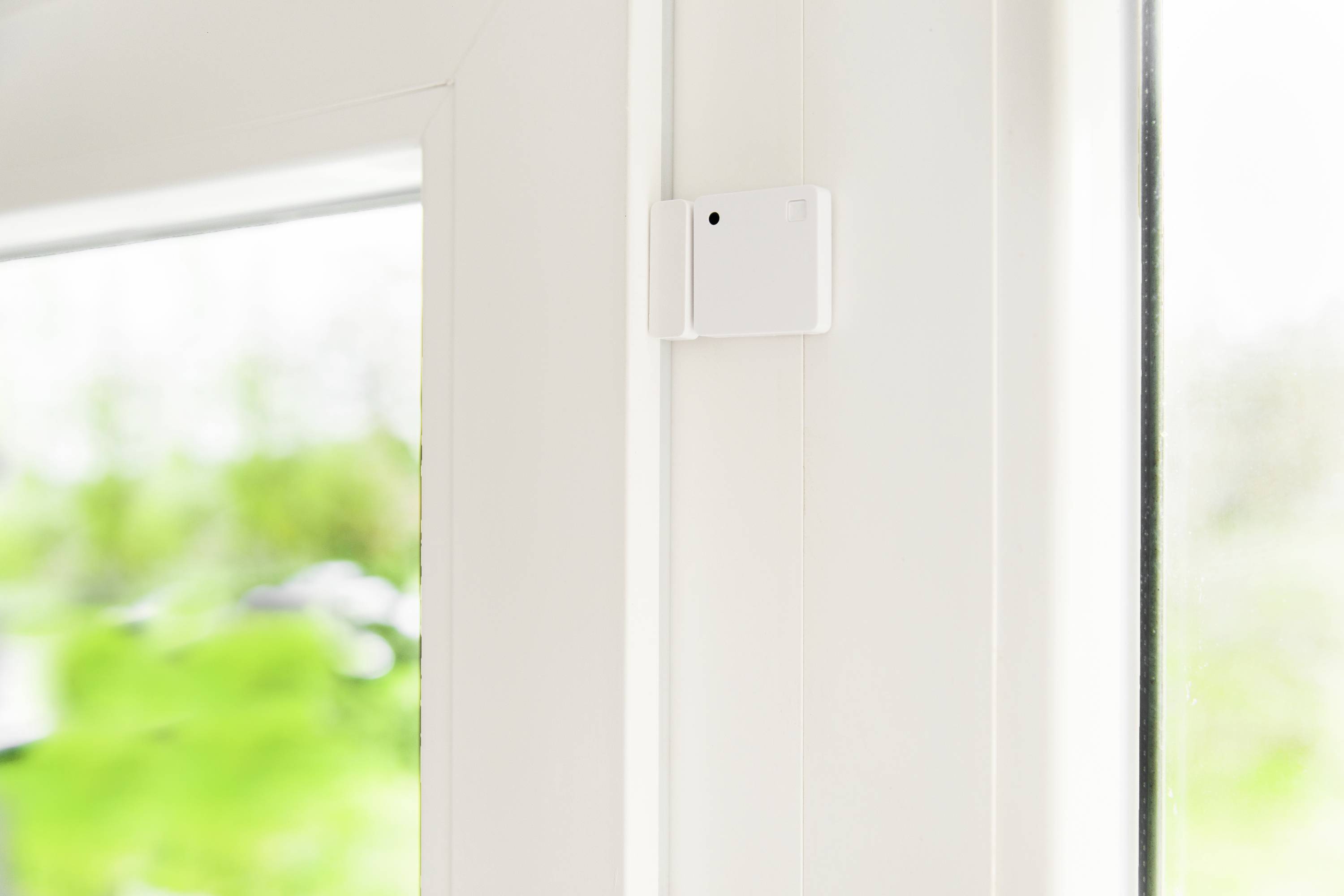 Ein weißer Fenstersensor ist oben auf dem Rahmen eines geöffneten Fensters montiert. Draußen sind verschwommene grüne Bäume sichtbar.