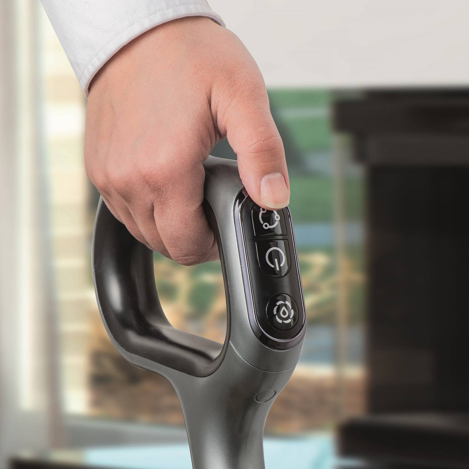 Eine Hand hält einen Controller eines Elektro-Rollstuhls, bereit zur Bedienung. Im Hintergrund sind verschwommene Wohnaccessoires erkennbar.