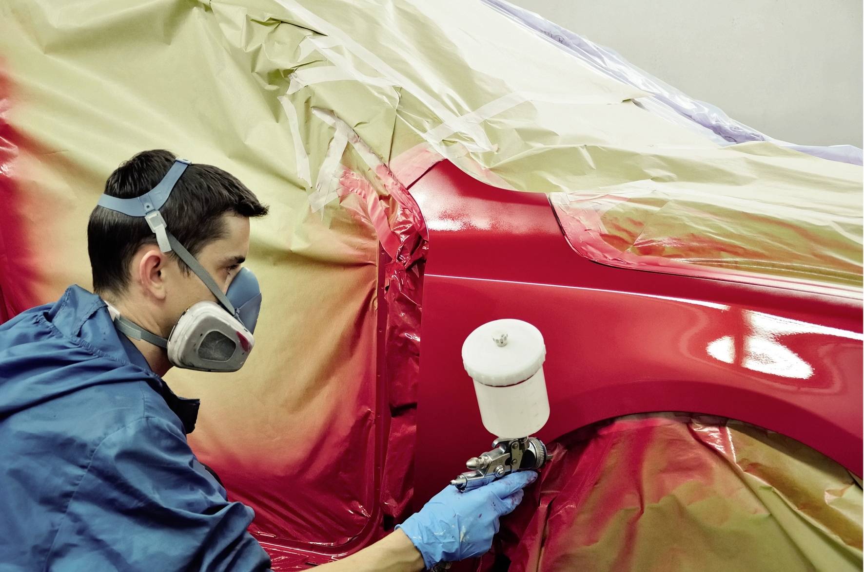 Ein Arbeiter sprüht rote Farbe auf ein Auto in einer Werkstatt. Der Mann trägt eine Schutzmaske und blaue Handschuhe.