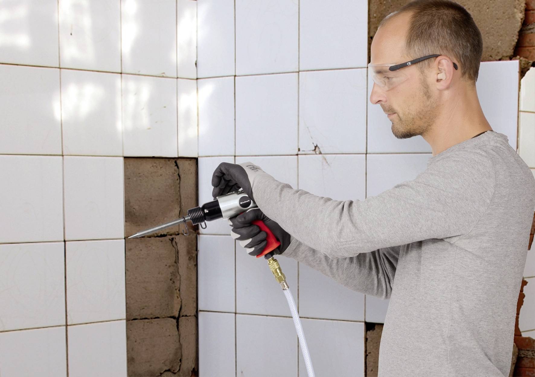 Ein Mann benutzt einen Drucklufthammer, um Fliesen an einer Wand zu entfernen. Schutzbrille und Handschuhe schützen ihn dabei.