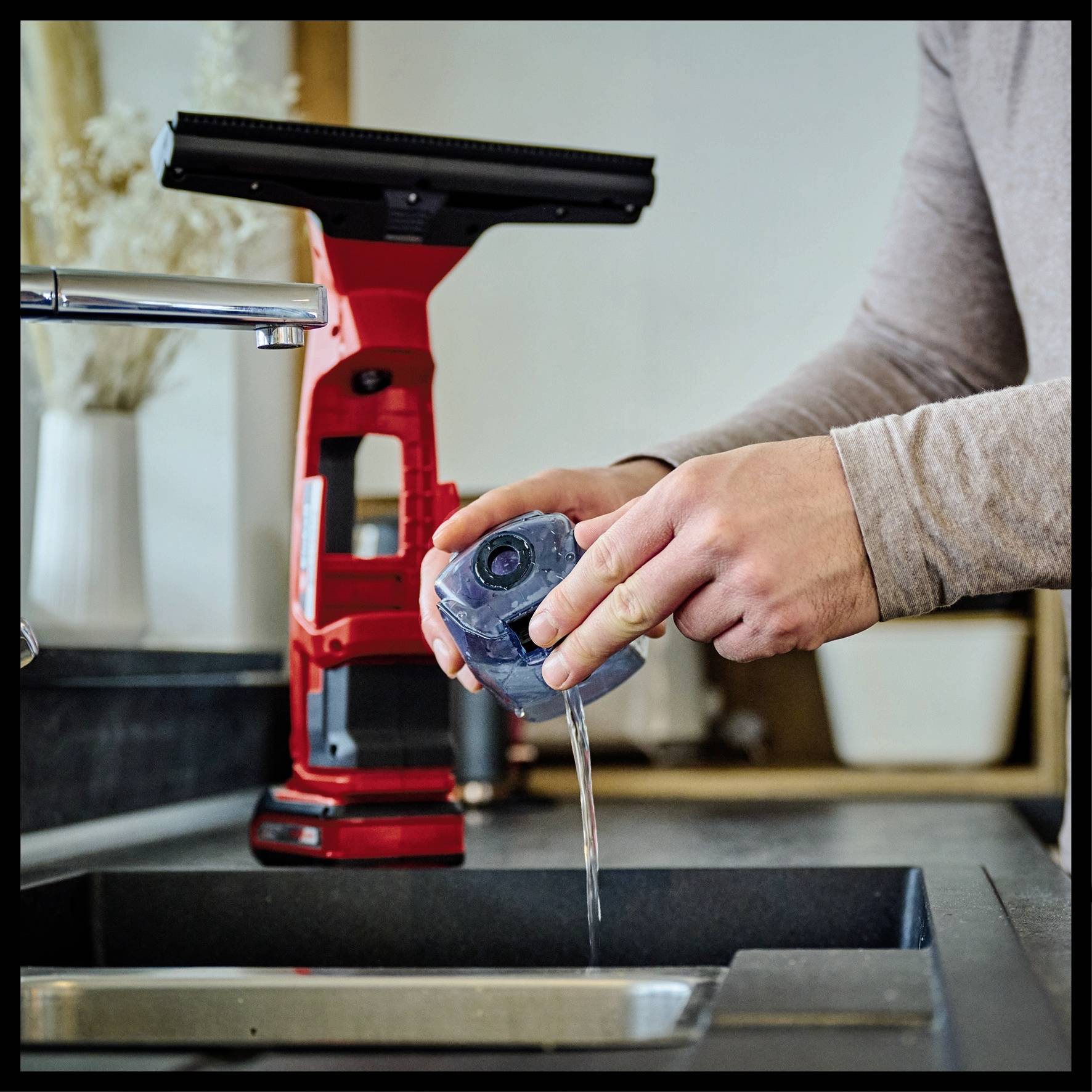 Eine Person füllt Wasser in einen Wassertank vor einem Wasserhahn. Im Hintergrund ist ein Fensterreiniger zu sehen.