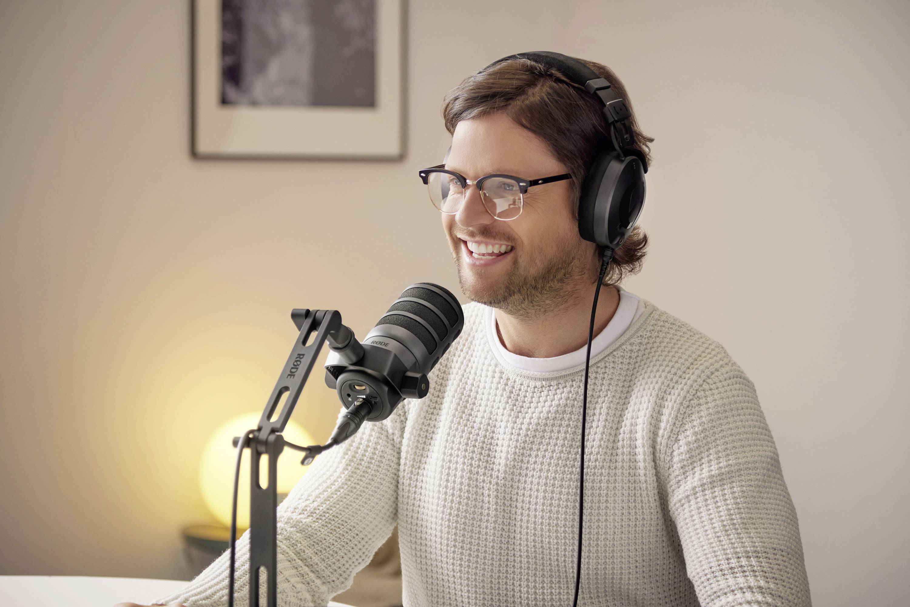 Person mit Kopfhörern und Mikrofon, sitzt lächelnd an einem Tisch, beteiligt sich an einer Audioaufnahme oder einem Podcast in einem gemütlichen Raum.