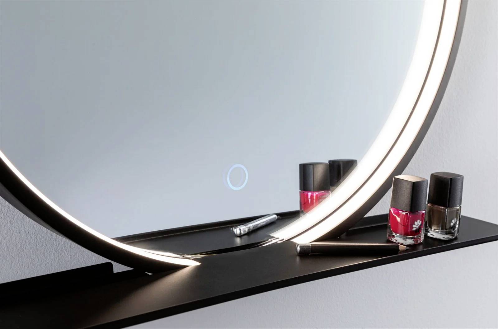 Ein runder Spiegel mit LED-Beleuchtung, dekoriert mit roten und schwarzen Nagellackflaschen auf einer Ablage darunter.