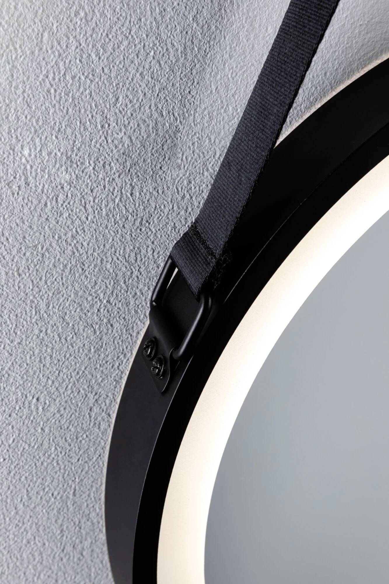 Nahaufnahme eines runden Spiegels an einer blauen Wand, aufgehängt mit einem schwarzen Gurt. Oben links im Bild sichtbar.